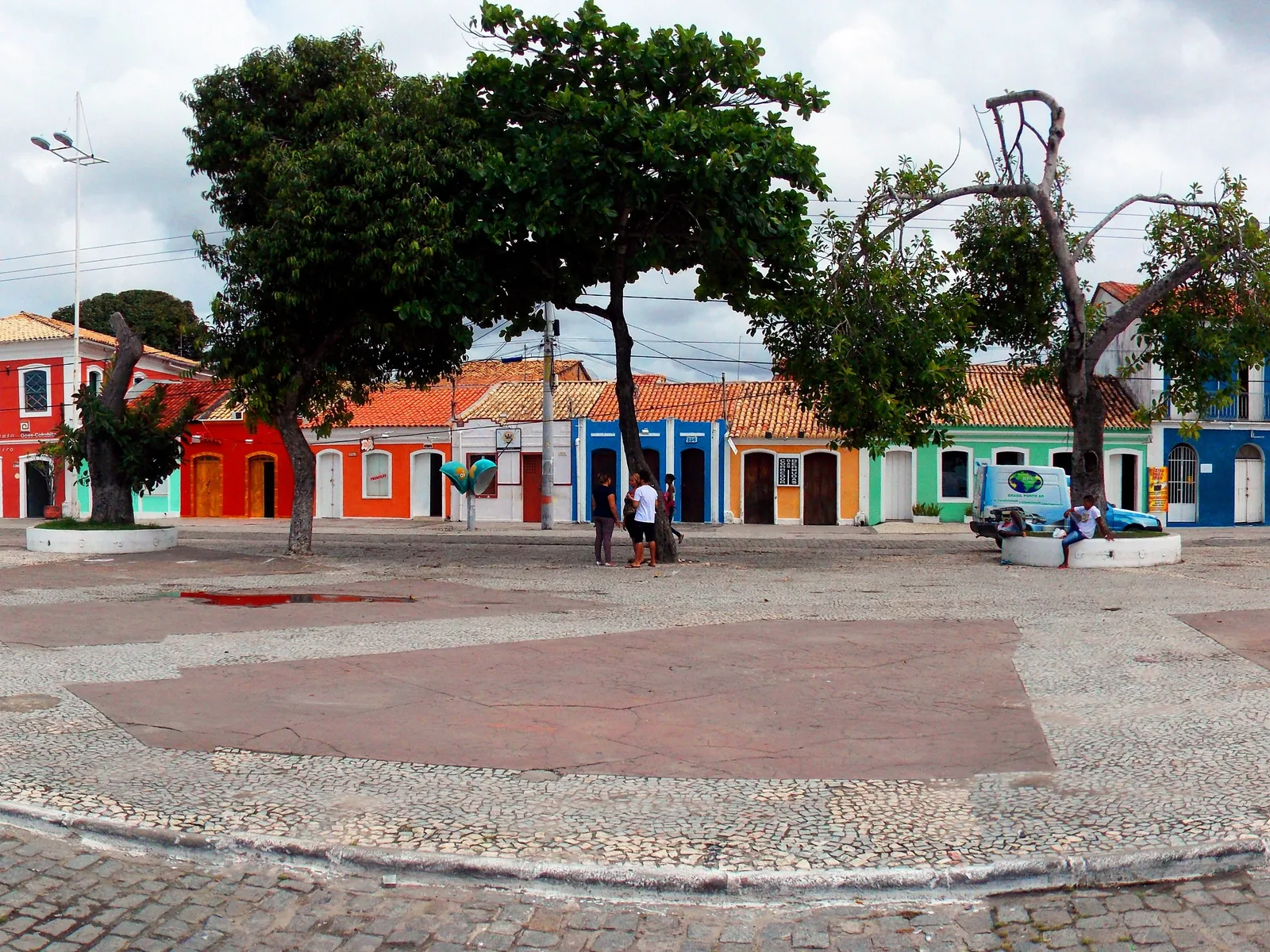 A wide paved square in the center of Porto Seguro by day, ringed by colorful low colonial buildings and shade trees, with a few people walking across it