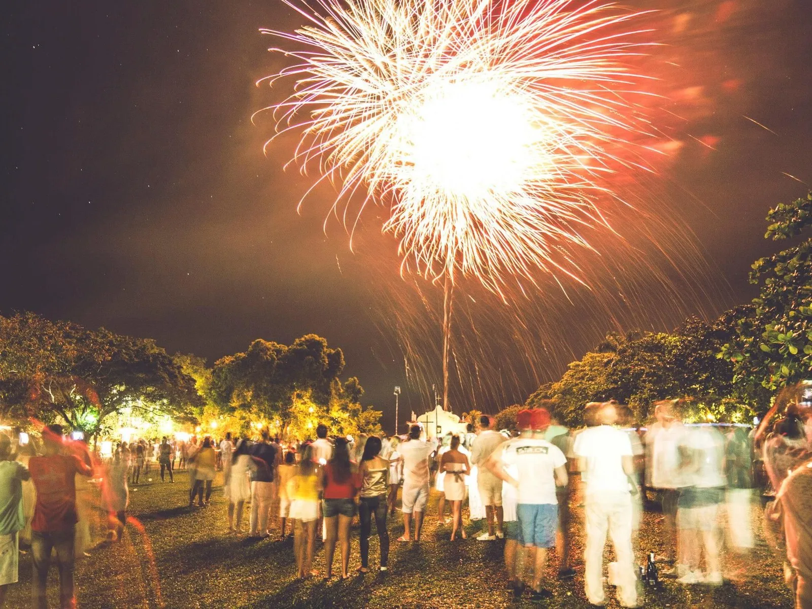 Fireworks bursting in gold and red over a crowd standing on the grass of Trancoso’s Quadrado on New Year’s Eve, lit trees behind them