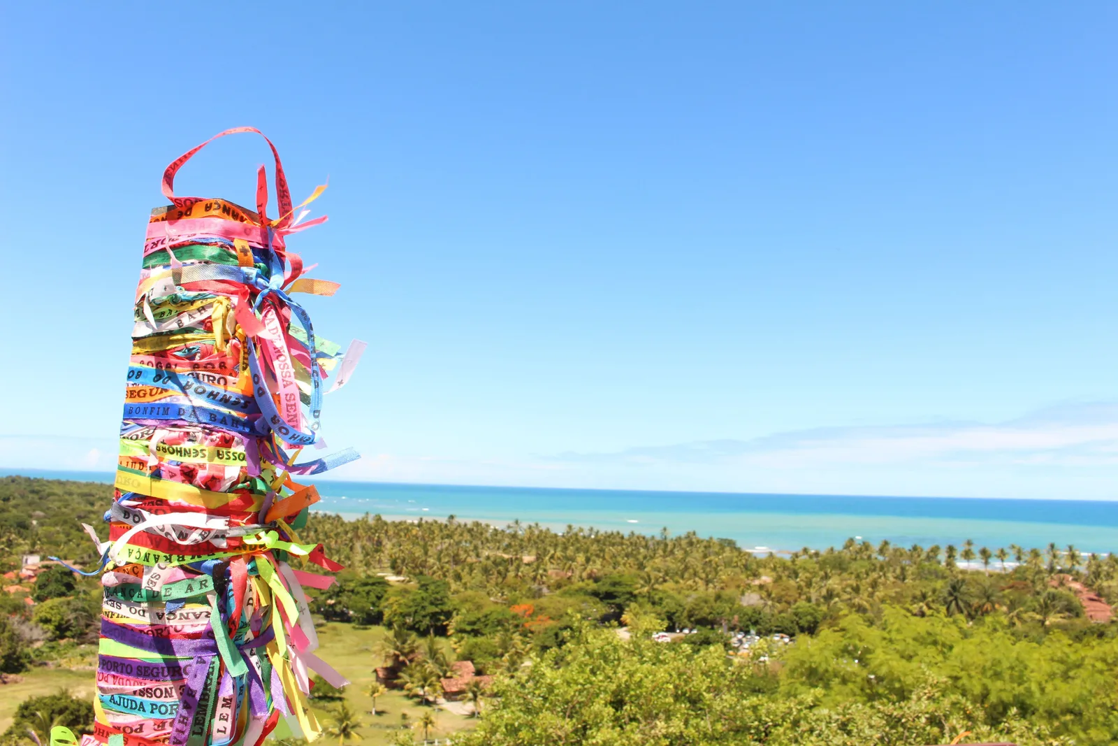 A post wrapped in hundreds of knotted colored ribbons by the church at Arraial d’Ajuda, with coconut palms below and the sea on the horizon