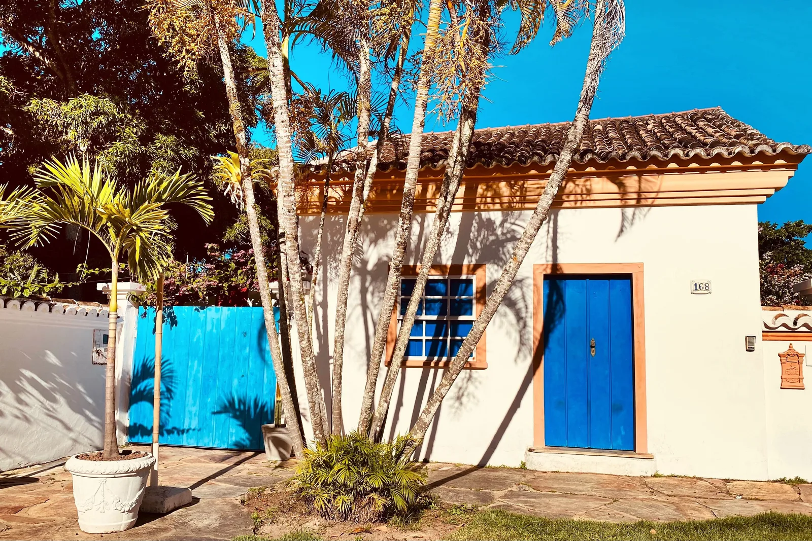 A whitewashed colonial house in Porto Seguro’s Cidade Alta with a blue door and a blue gate beside it, thin palms growing in front