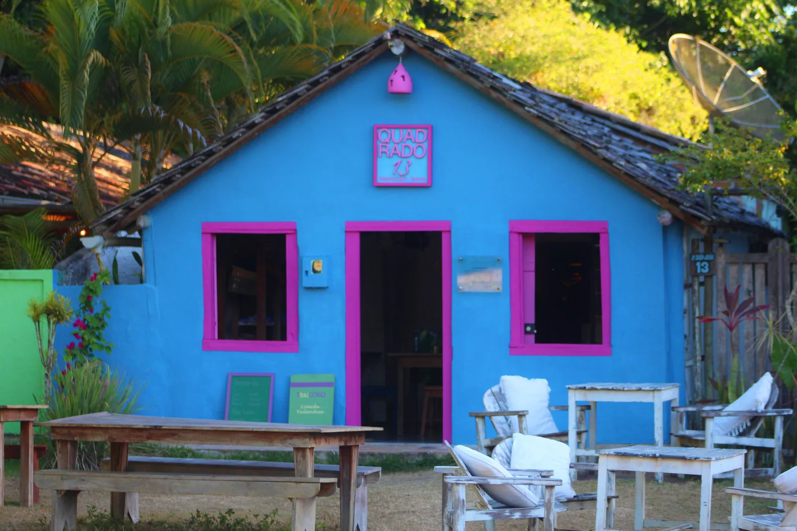 A bright blue cottage on Trancoso’s Quadrado with hot-pink window and door frames, a green wall beside it, and white chairs and long wooden tables set out in front