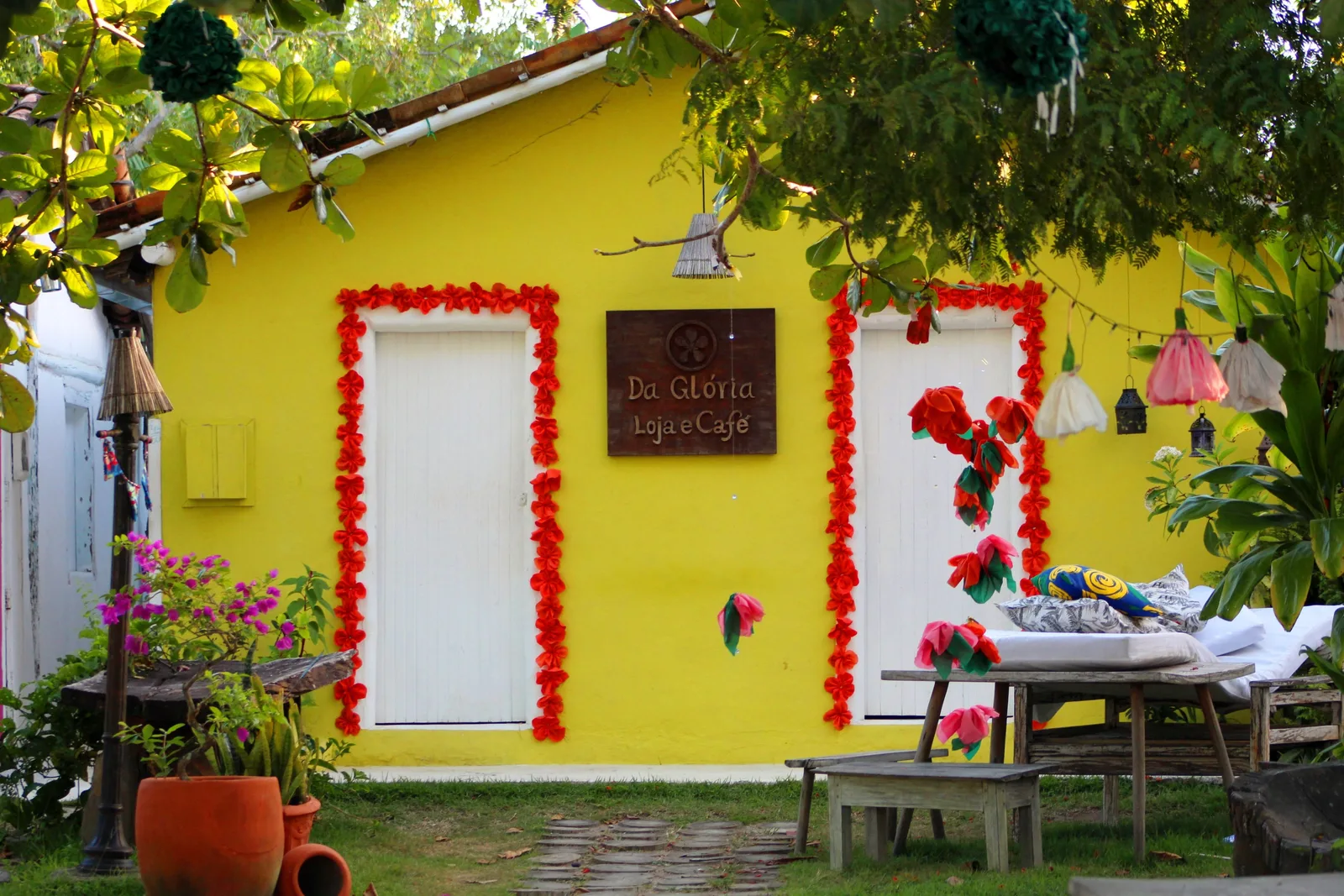 A yellow cottage on the Quadrado with two white doors framed in garlands of red flowers, potted plants, hanging lanterns and a table set out beside it