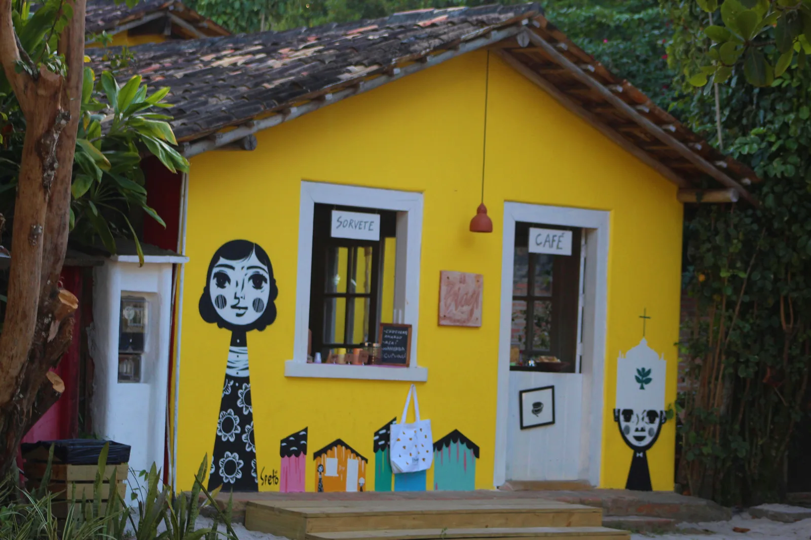 A yellow cottage on Trancoso’s Quadrado with two large folk-art faces and a row of little houses painted on its front wall, a white-framed window and a white door