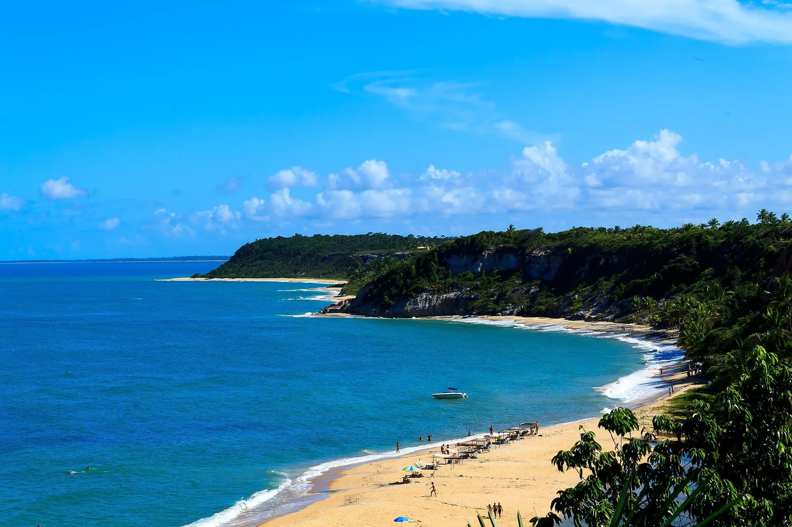 A beach near Trancoso from above, a line of thatched shades and a few people on the sand, one white motorboat anchored offshore and forested bluffs with pale cliffs running out to a headland