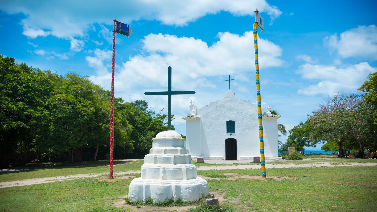 The small whitewashed church at the end of Trancoso’s Quadrado, a dark cross on a stepped white plinth in front of it and two tall striped poles with flags either side, mown grass and trees around it and a sliver of sea at the right edge