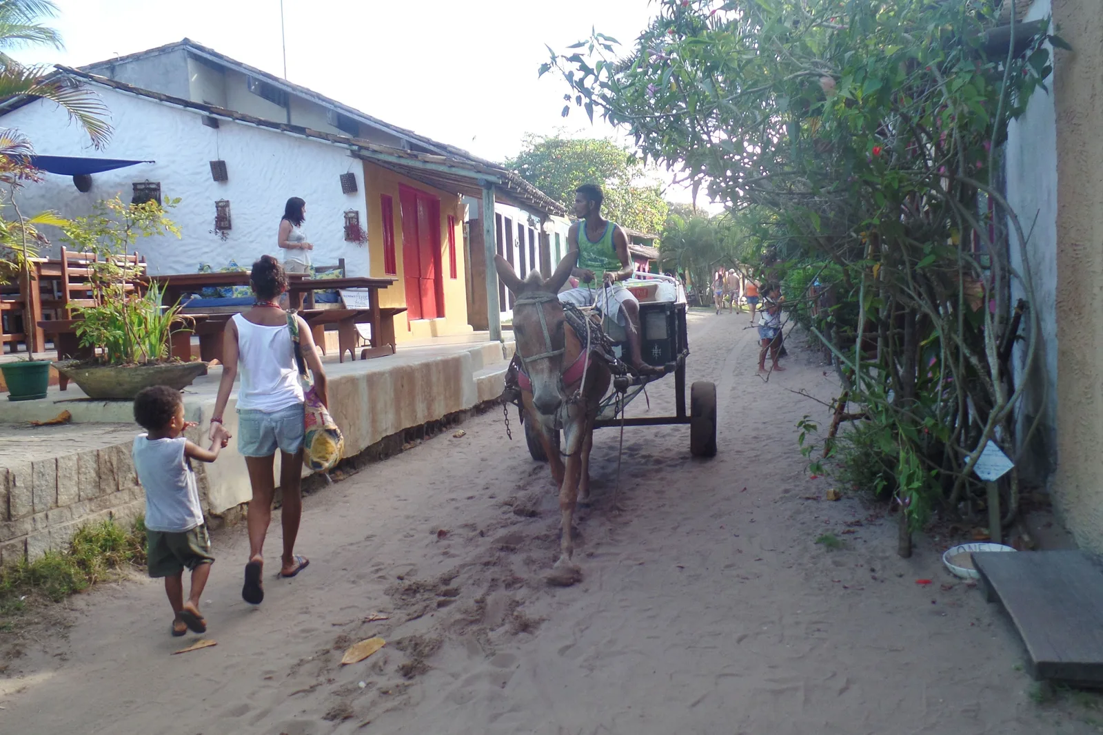 A sand street in Caraíva: a woman and a small boy walking past a mule pulling a cart with a man aboard, a whitewashed house with red doors on one side and bushes on the other
