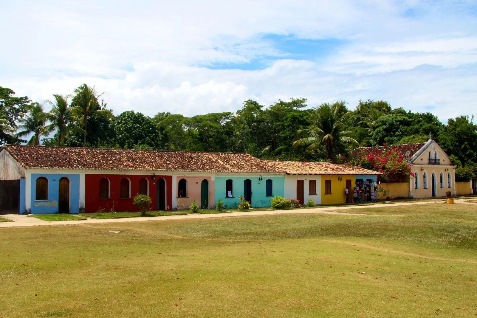 A row of low colonial houses painted blue, red, yellow and white across a wide lawn in Porto Seguro’s Cidade Alta, trees rising behind them