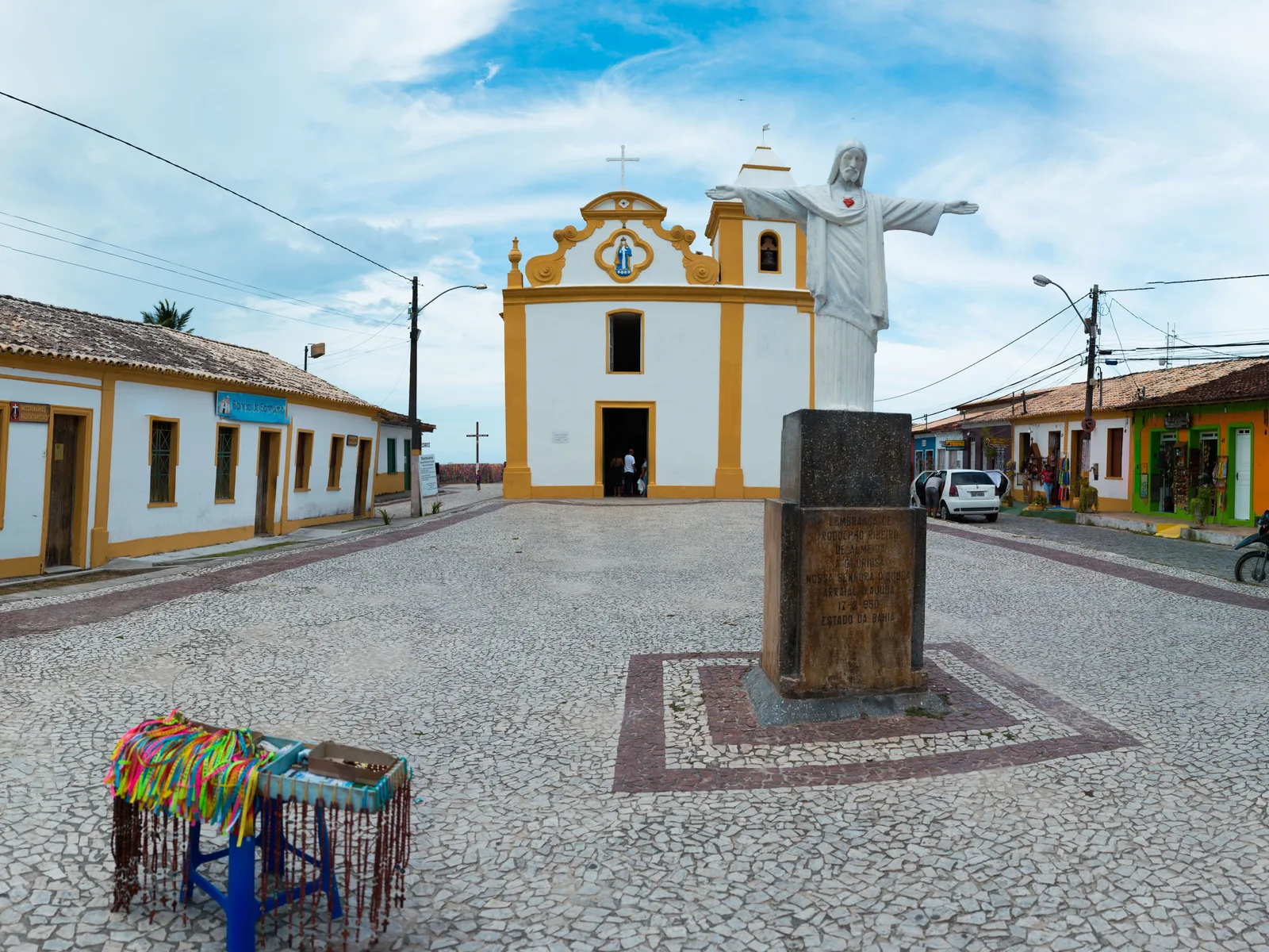 The white and yellow church of Nossa Senhora d’Ajuda at the end of a paved square in Arraial d’Ajuda, a white statue of Christ with outstretched arms on a dark stone pedestal in front and low painted houses on both sides