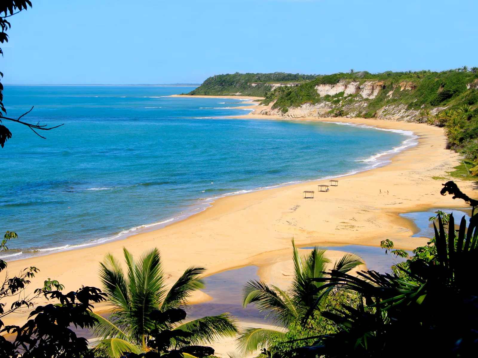 Praia do Espelho framed by dark foliage: a curve of pale sand along turquoise water, low reddish cliffs with forest on the headland beyond, and a stream crossing the beach in the foreground