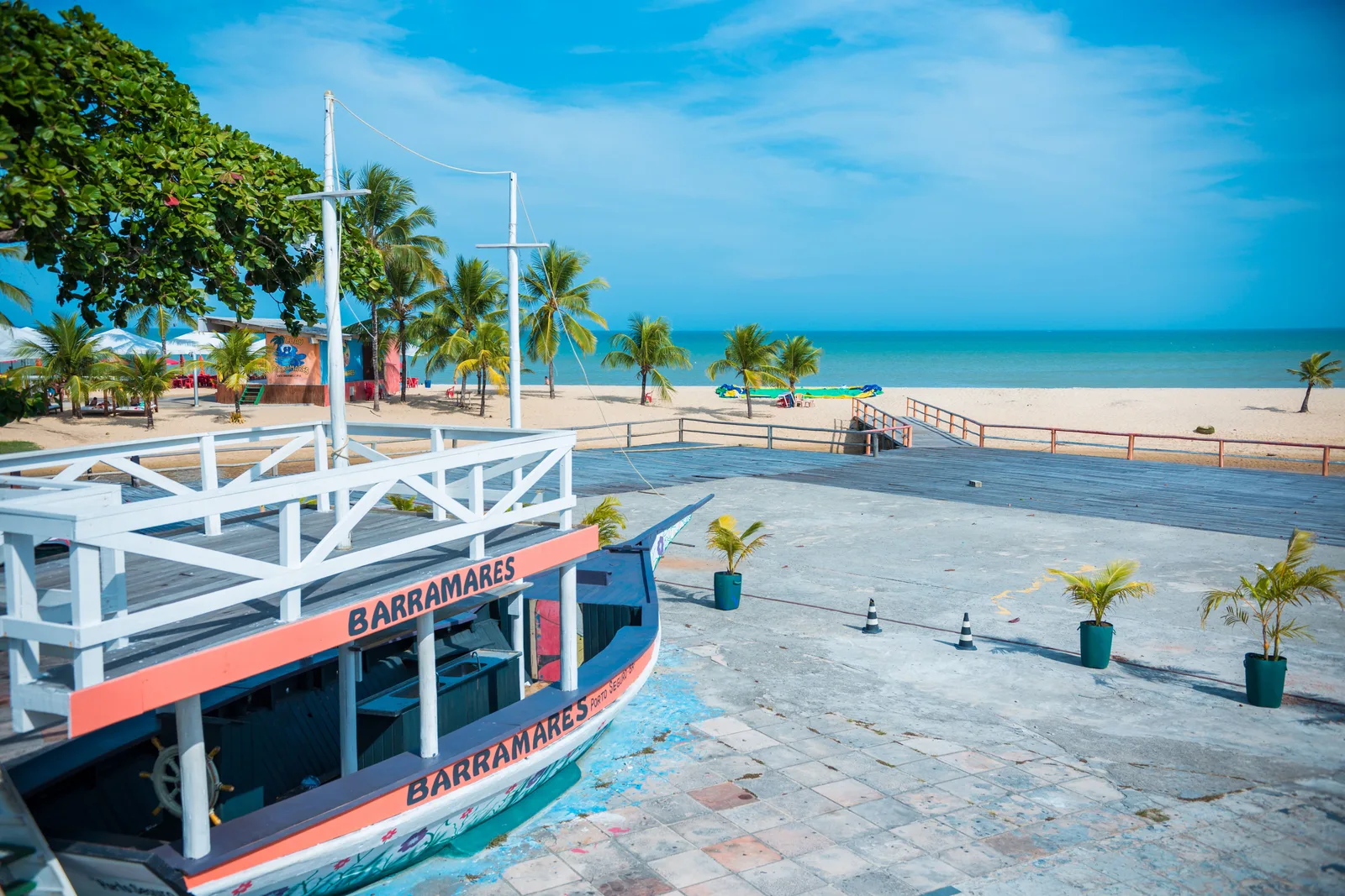 A navy boat with an orange-red band and the name Barramares, white rails and two masts, standing on a paved beach terrace in Porto Seguro, potted palms around it and the beach and sea behind