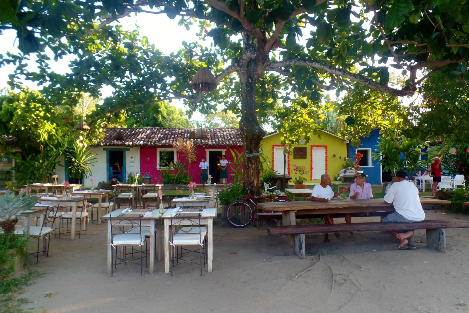 Tables and benches set out on sandy ground under a big shade tree on Trancoso’s Quadrado, a few people sitting at them, a bicycle, and red, yellow and blue painted cottages behind