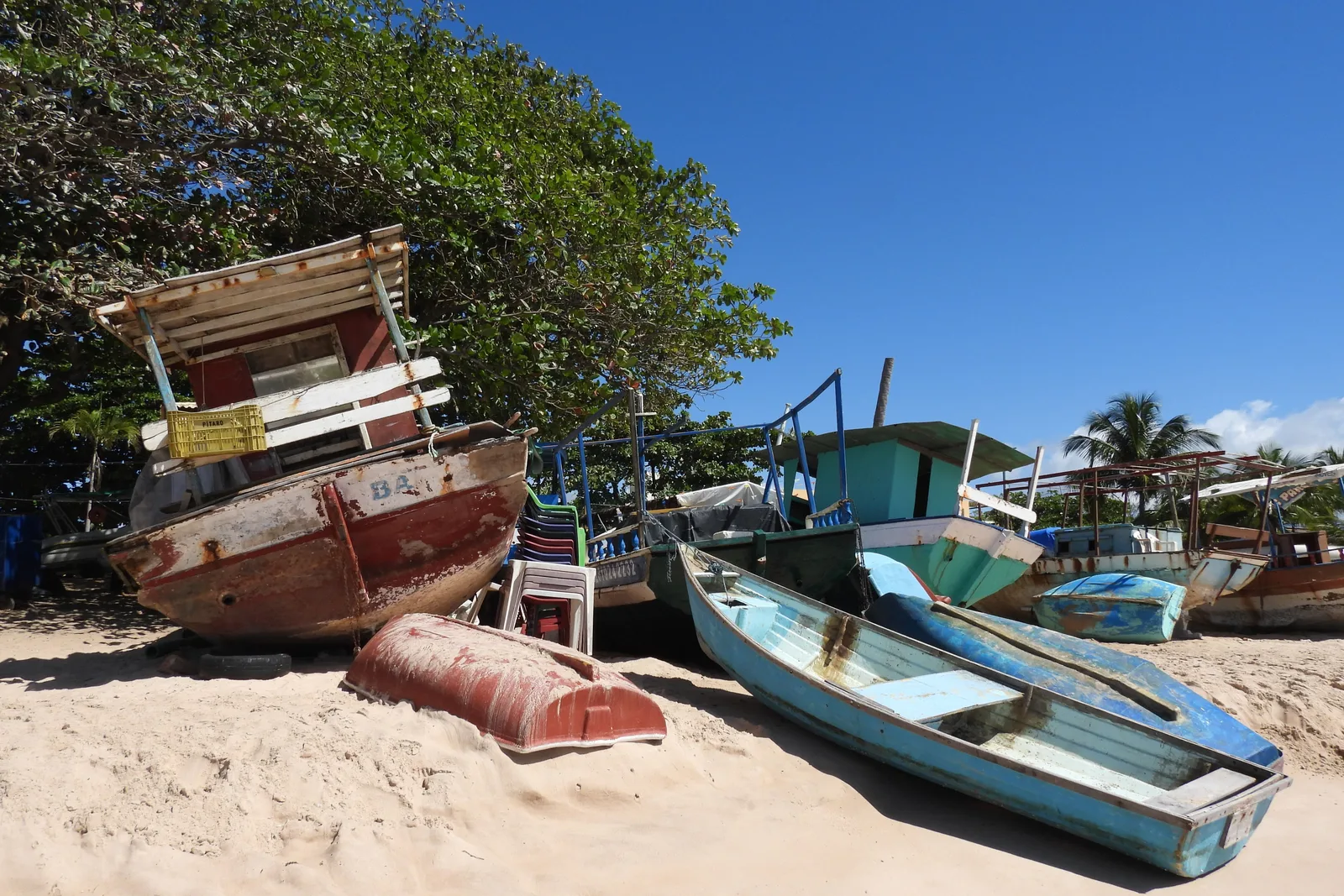 Painted wooden fishing boats pulled up under the trees on the beach at Itacimirim