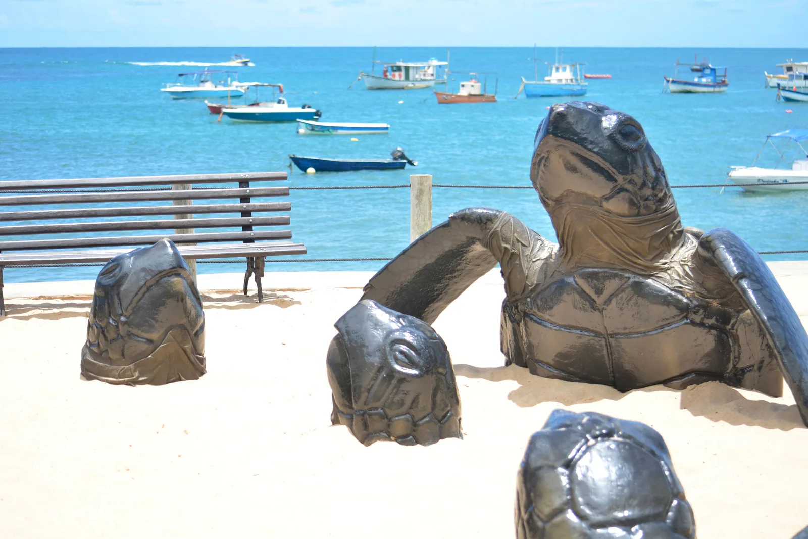 Metal sea turtle sculptures on the sand by the Projeto Tamar center, fishing boats moored beyond