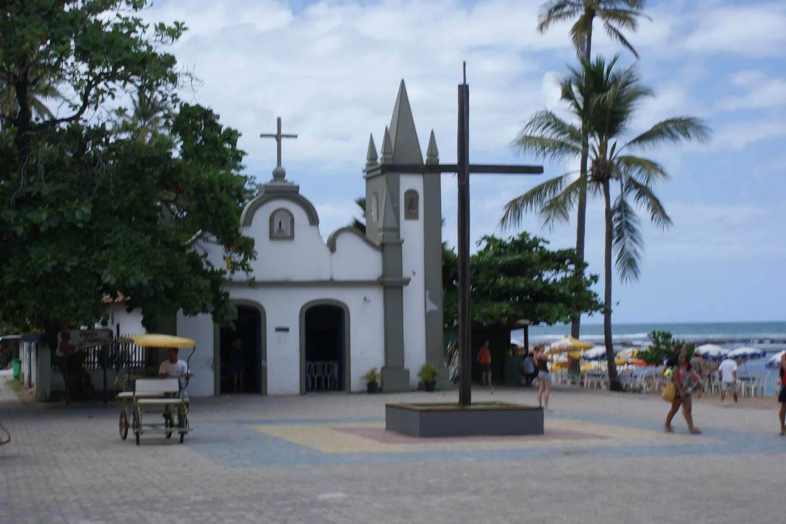 The small white chapel on the square in Praia do Forte village, with a tall cross, palms and the sea behind