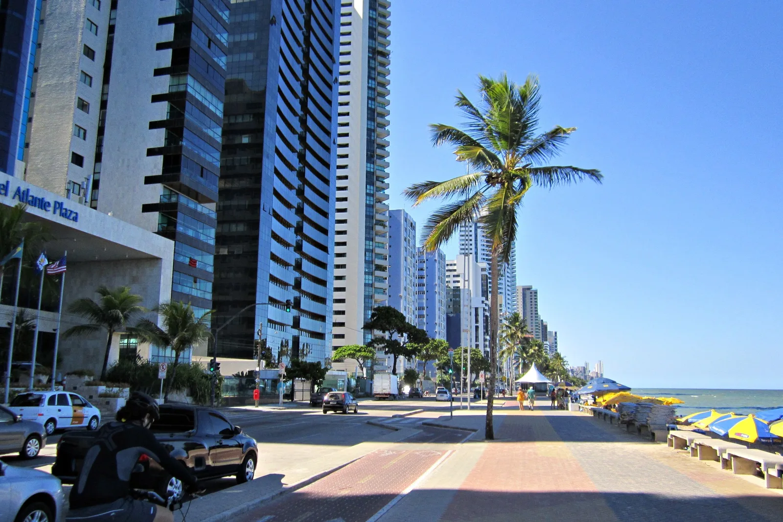 The palm-lined promenade and red bike lane along the Boa Viagem seafront in Recife, apartment towers on one side and beach umbrellas on the sand at the other