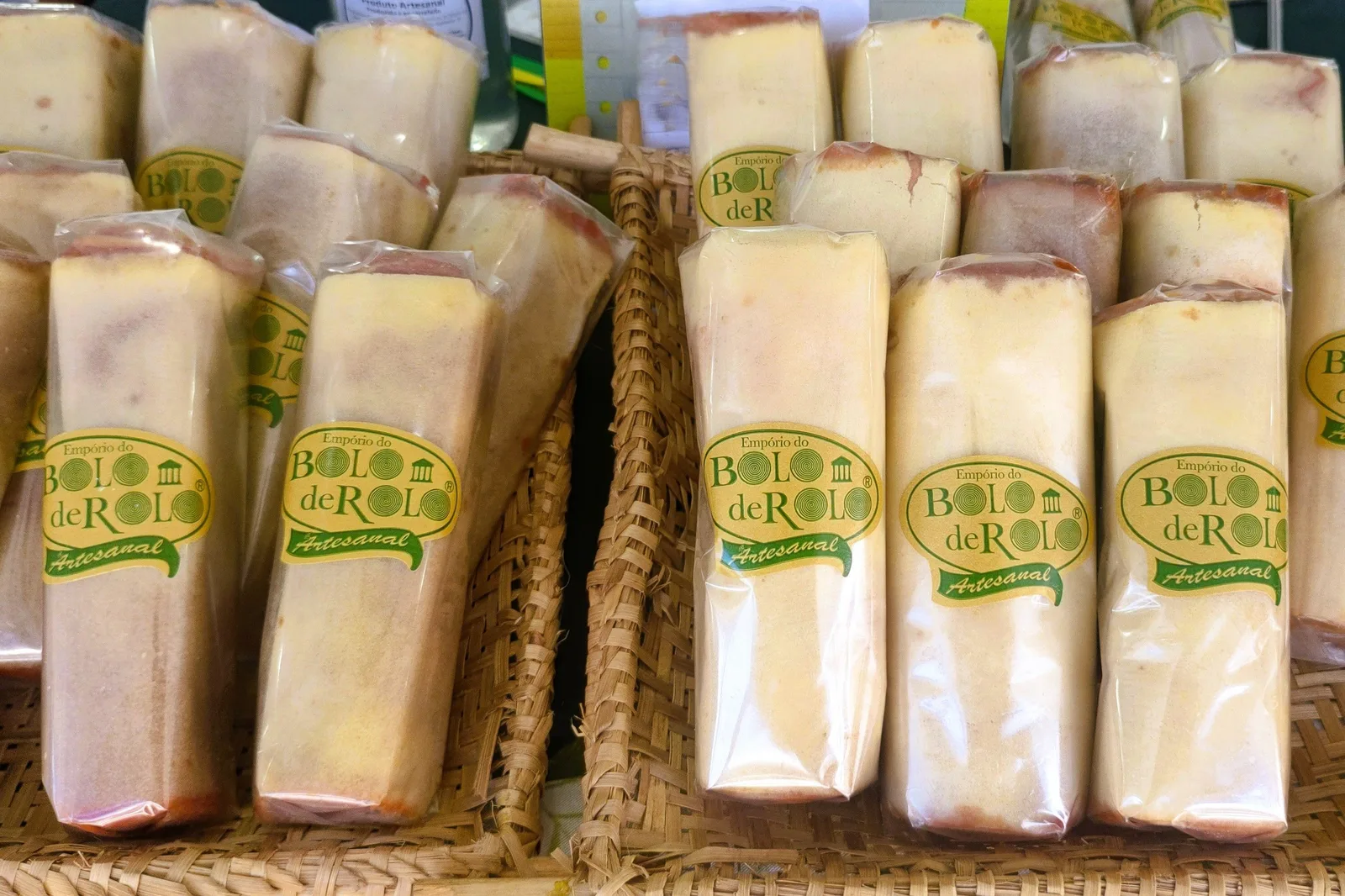 Cellophane-wrapped bricks of bolo de rolo standing on end in a woven basket at a market stall, green and gold labels facing out and a seam of guava showing along the top of each