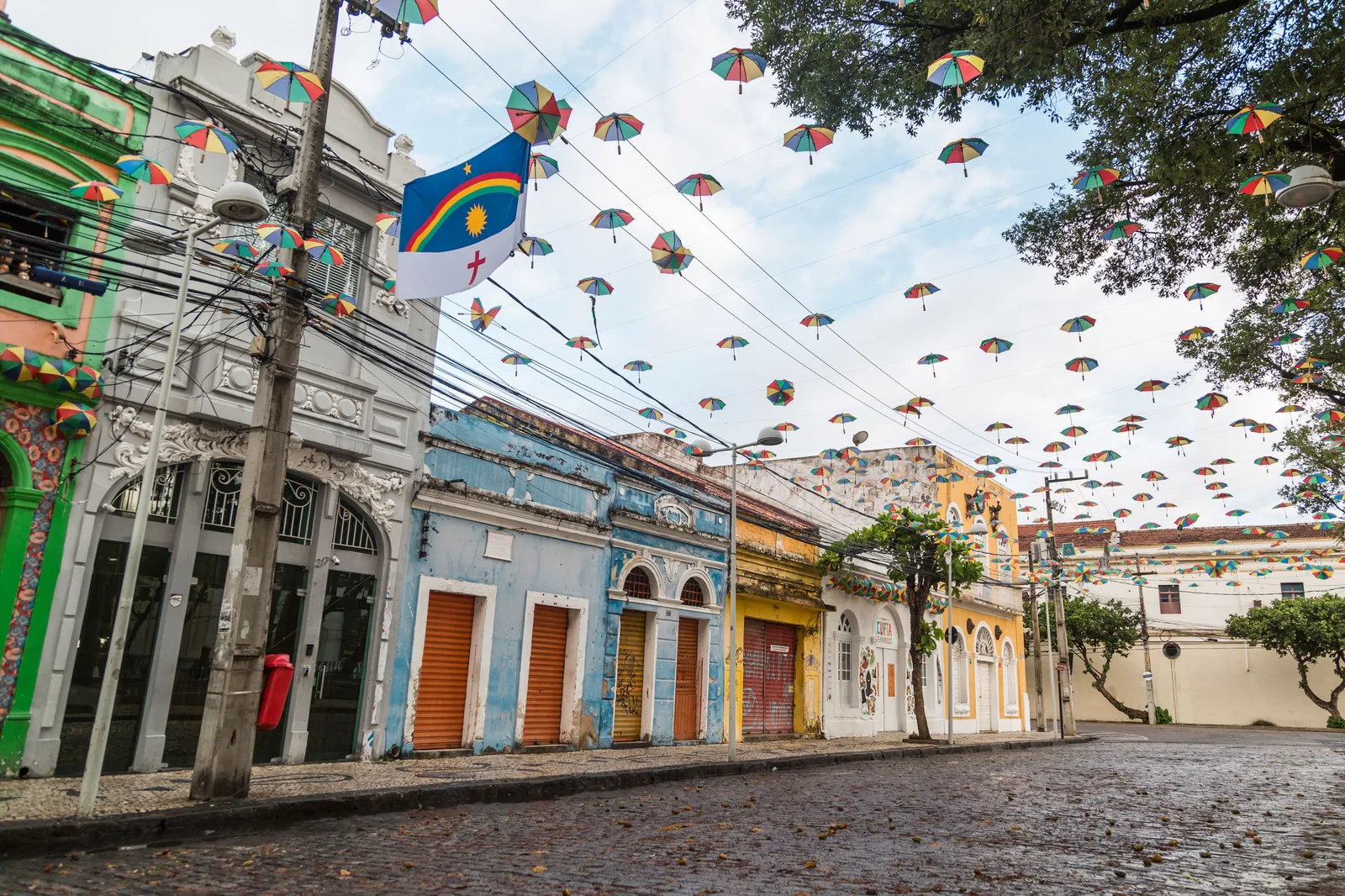 A street of single-story shops in Recife with orange roller shutters and painted fronts, hundreds of small carnival umbrellas strung on wires overhead and a Pernambuco flag flying