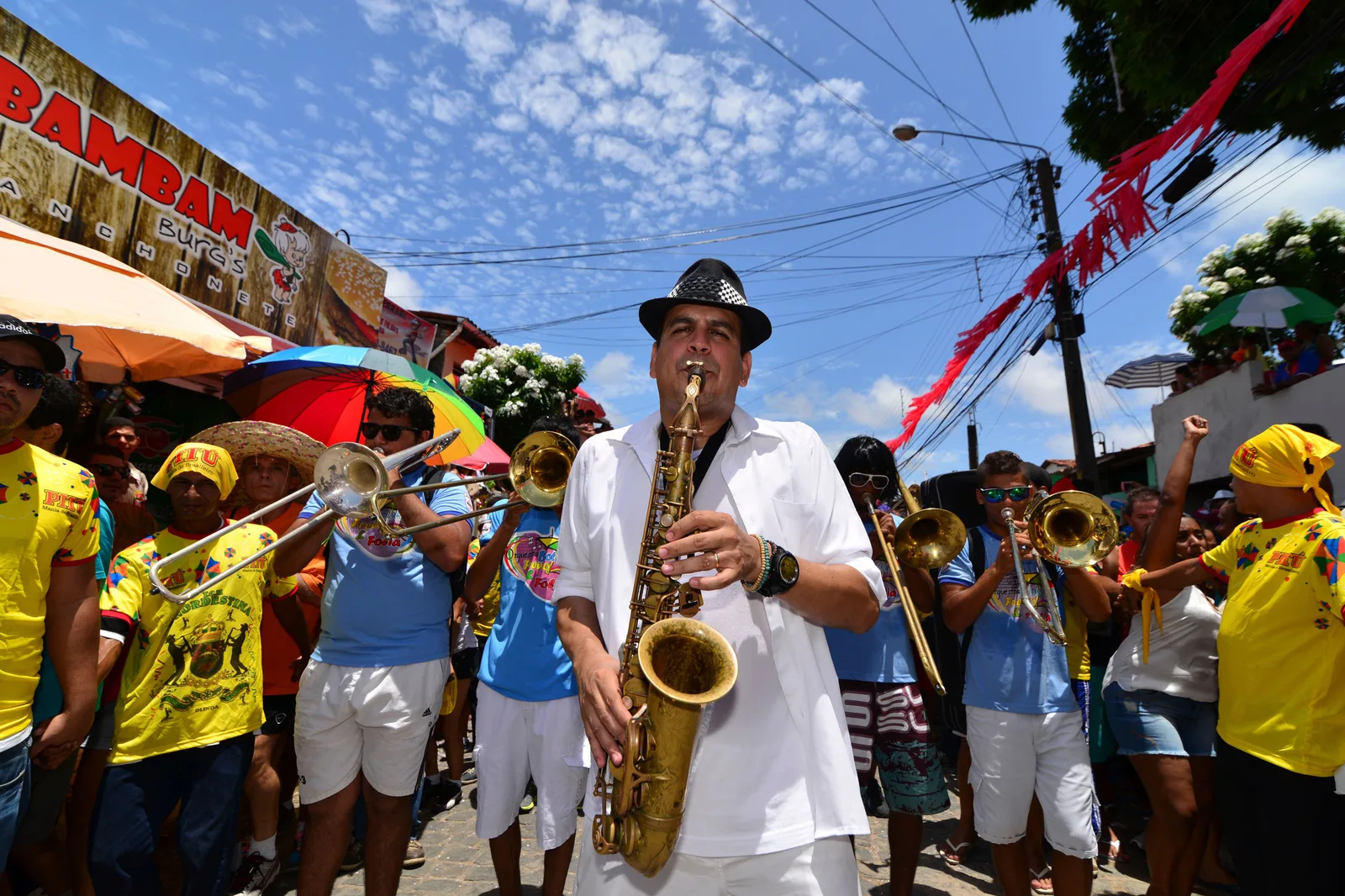 A carnival brass band moving down a street hung with streamers, a saxophonist in white at the front and trumpet and trombone players behind him in yellow and light blue shirts