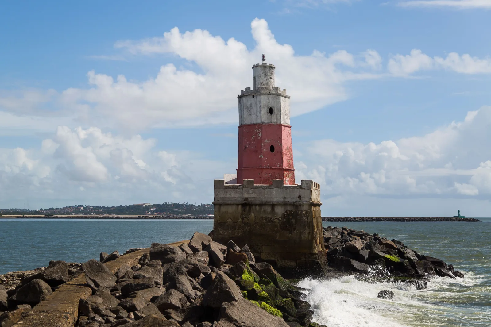 The red and white lighthouse of Recife standing on the rocky stone reef at the harbor mouth, with surf breaking against the boulders below it