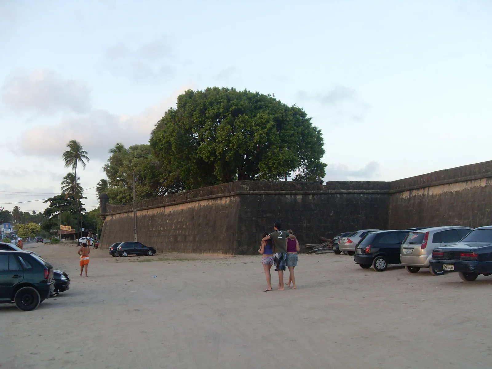 The low brick and stone rampart of Forte Orange on Itamaracá under a large shade tree, with visitors walking along the open ground in front of it