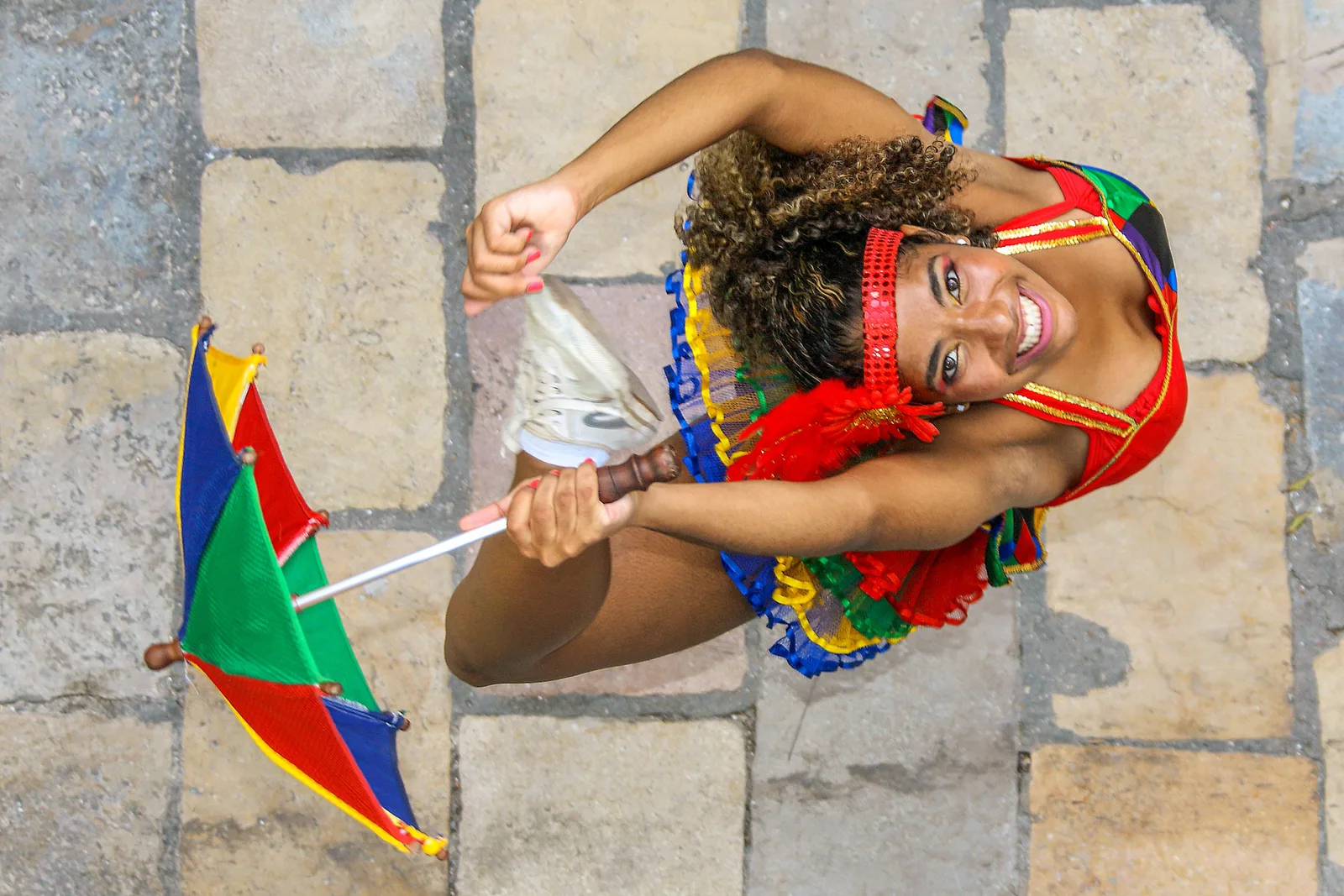 A frevo dancer photographed from above on cobblestones, in a red, green and yellow costume, laughing and holding a small brightly colored umbrella