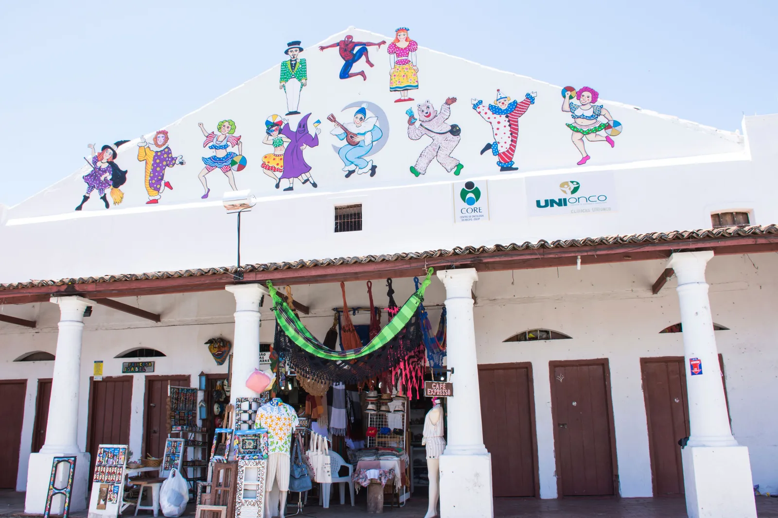 The white colonnaded front of the Mercado da Ribeira in Olinda, its pediment painted with dancing carnival figures and craft stalls set up under the arcade