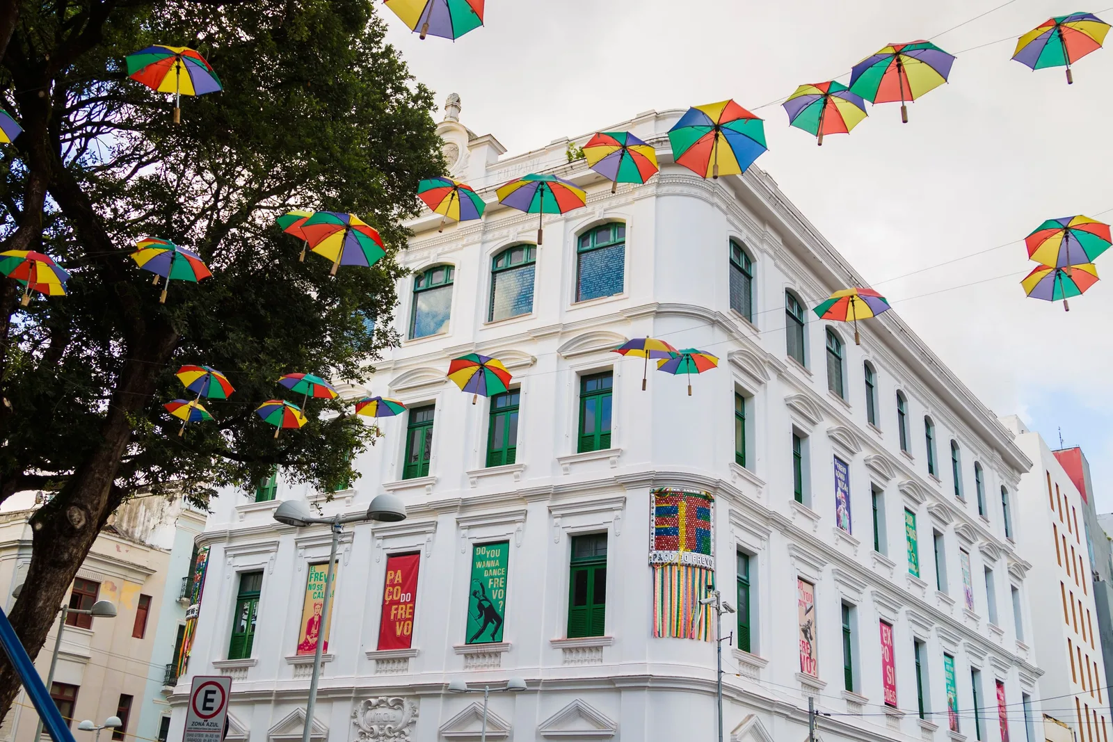 The white colonial facade of the Paço do Frevo in Recife Antigo, with rows of small colored carnival umbrellas strung on wires overhead against a blue sky