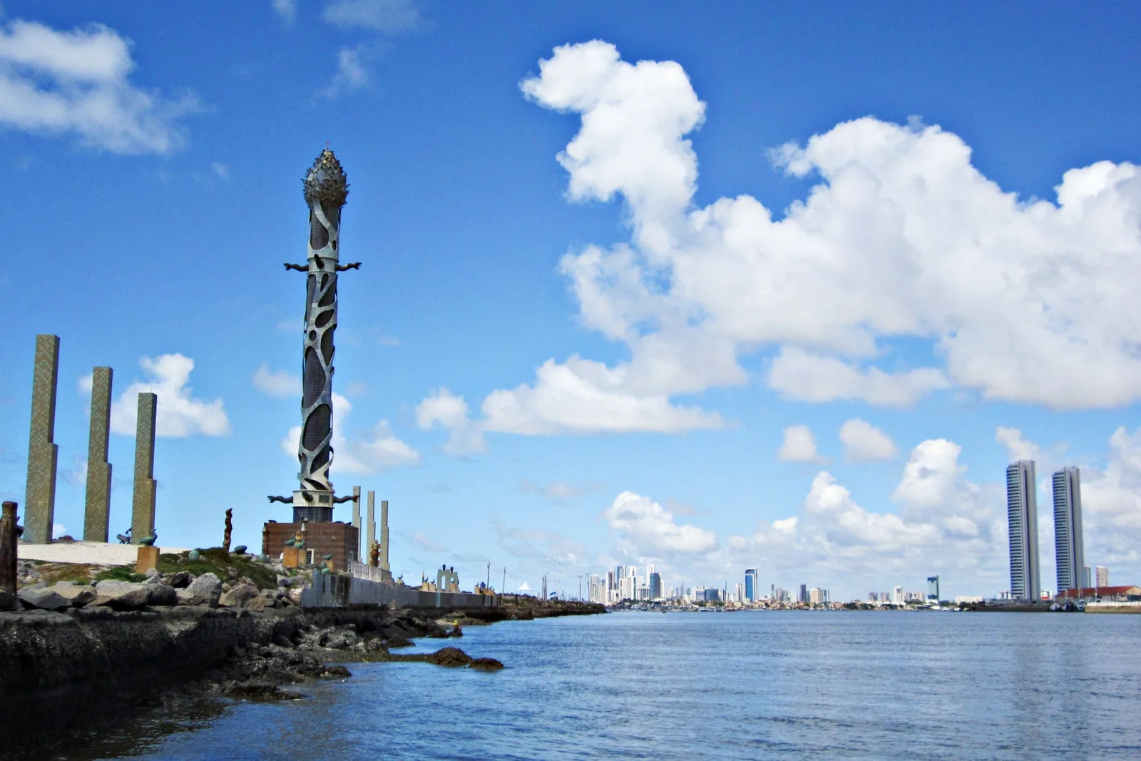 A tall glazed ceramic sculpture standing on the stone breakwater at Recife with the sea around it and the city’s skyline low on the horizon behind