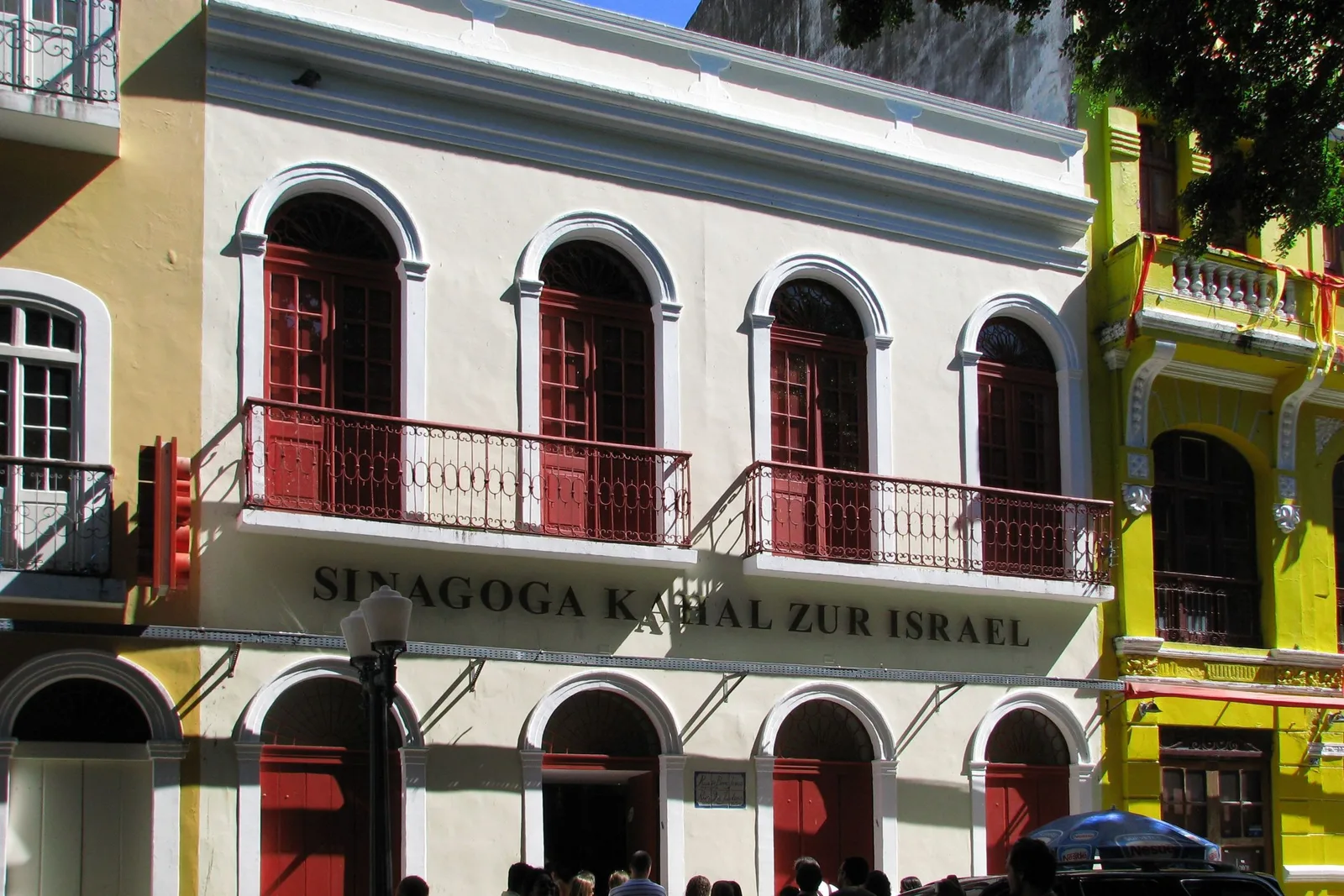 The restored front of the Kahal Zur Israel synagogue on the Rua do Bom Jesus: a cream facade with red-shuttered arched windows and dark red ironwork balconies, its name lettered across the middle, and a bright yellow neighbor beside it