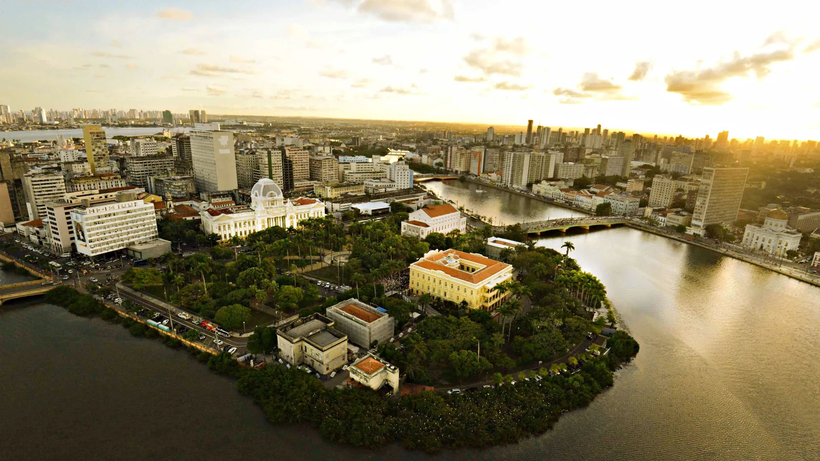 Recife from the air at golden hour, the Capibaribe curving around a wooded point with the governor’s palace on it and the city stretching away behind