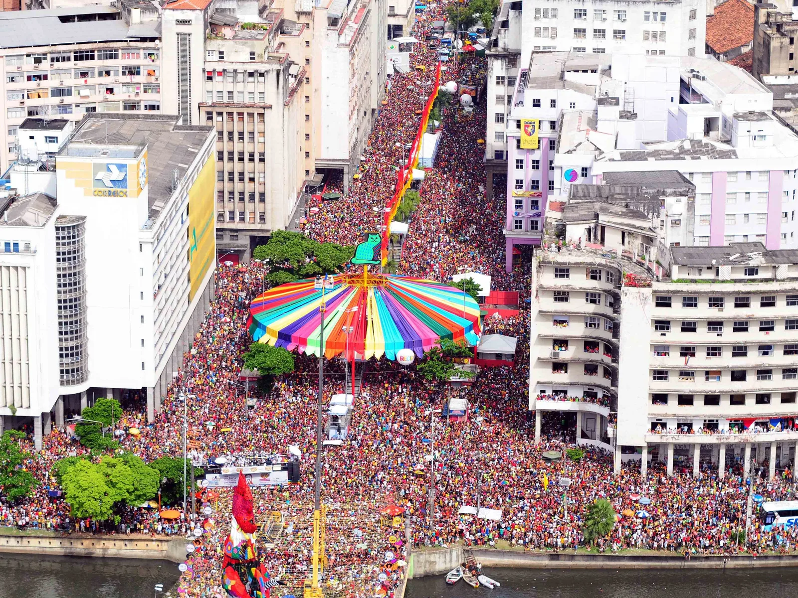 An aerial view of a wide avenue in central Recife filled curb to curb with a carnival crowd, a giant rooster float standing over it beside an enormous multicolored umbrella