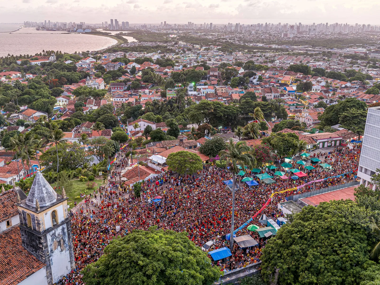 Olinda from the air during carnival: a church tower in the foreground and the streets below it packed solid with a crowd stretching down the hill toward the sea