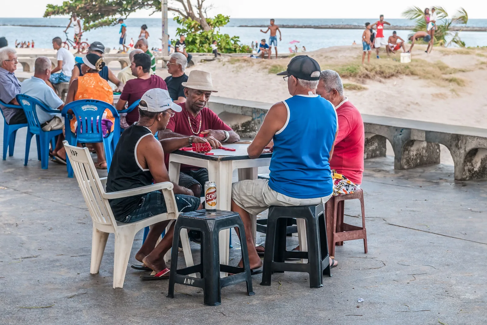Men playing dominoes at a plastic table on the seafront at Olinda, with the beach and the water behind them