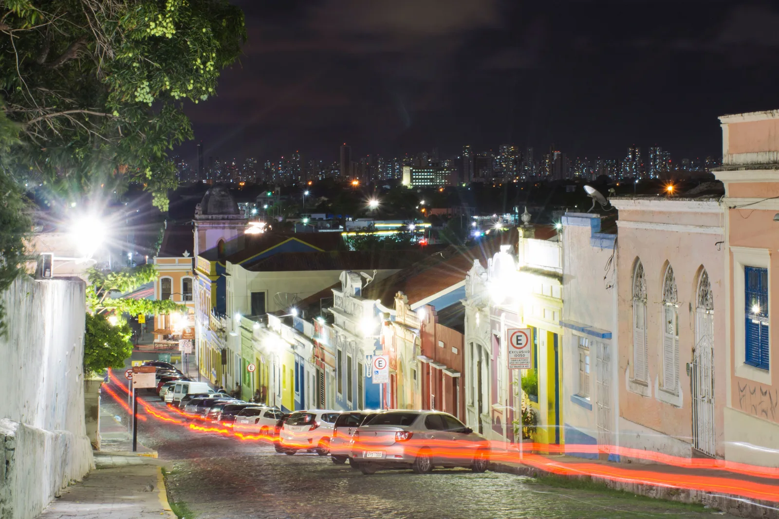 A street in Olinda’s historic center at night, its colonial facades lit and the lights of Recife strung along the horizon behind