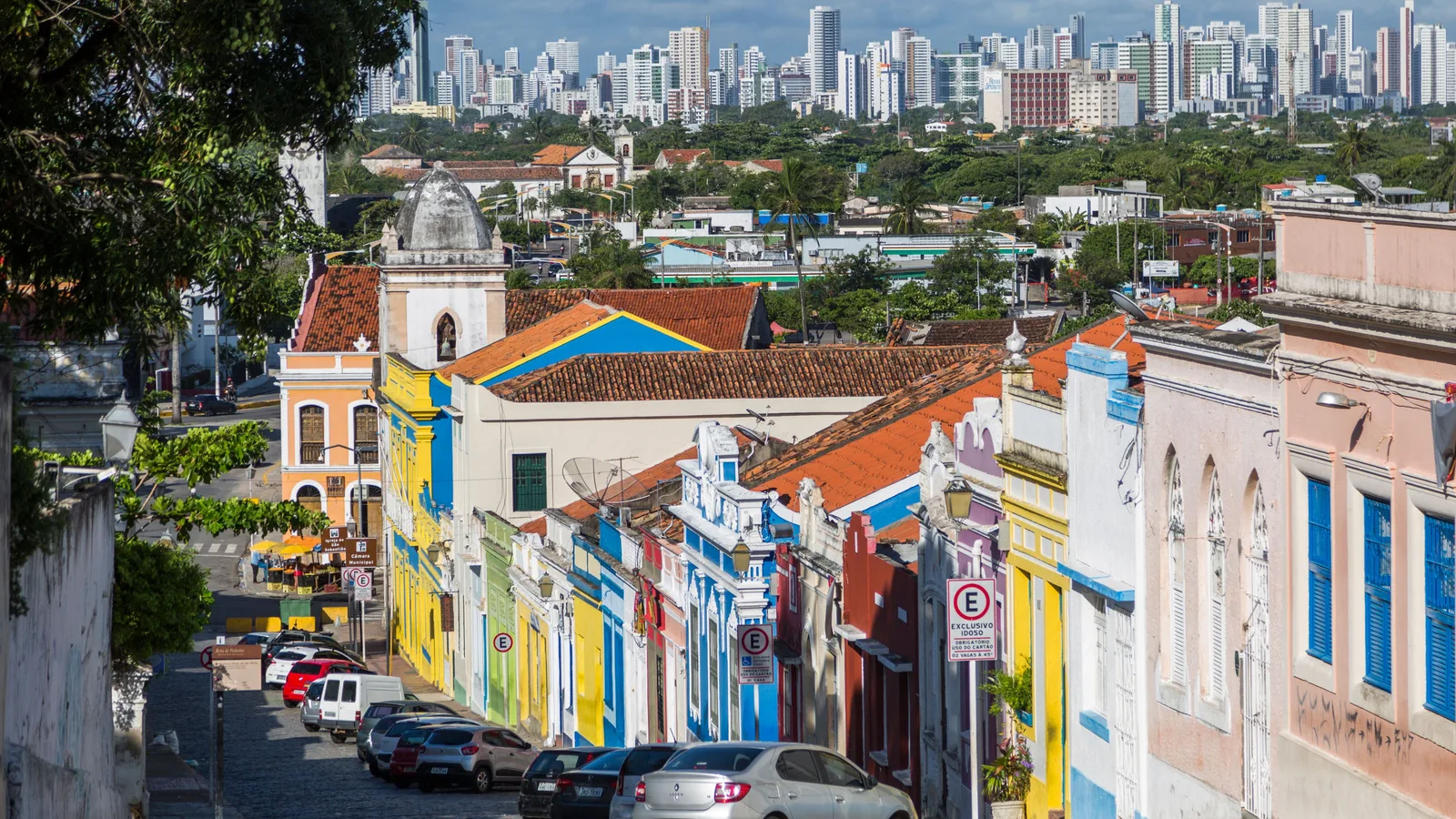 A steep street in Olinda's historic center lined with houses painted yellow, blue, pink and green under terracotta roofs, with the high-rise skyline of Recife stretched across the horizon beyond