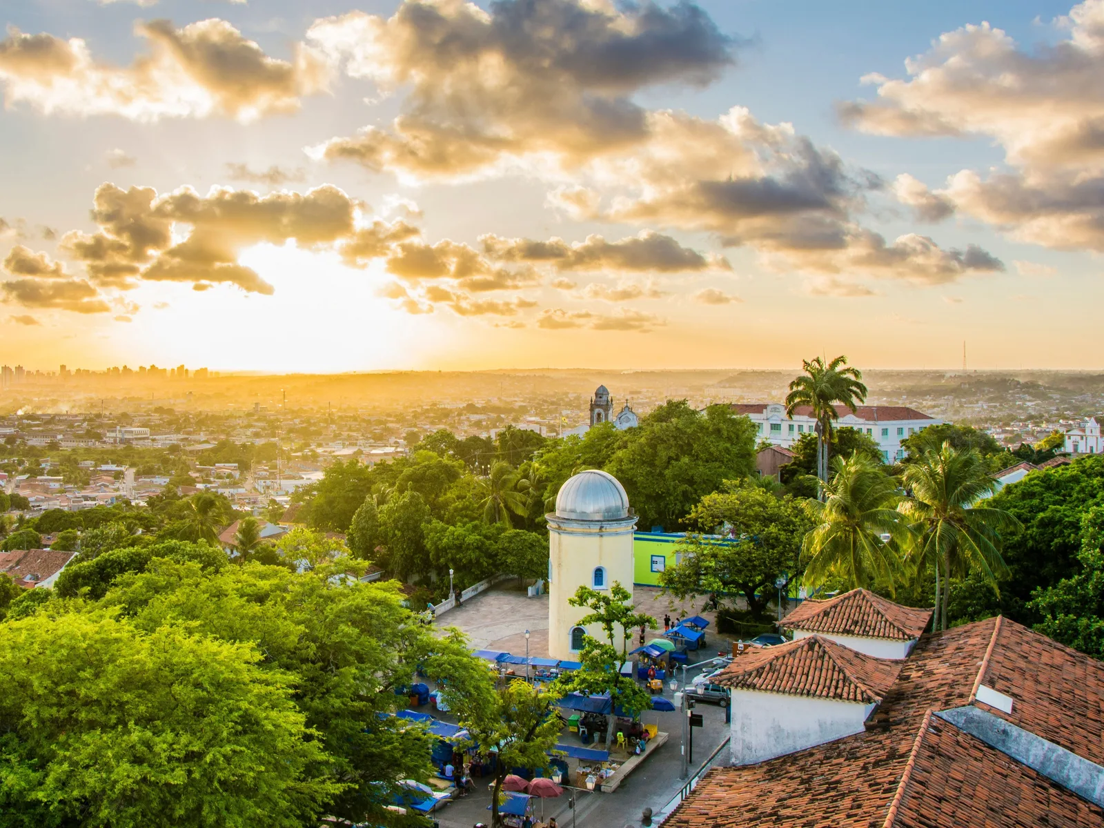 Golden light over Olinda at the end of the day: the silver dome of the Alto da Sé water tower and terracotta roofs among dense trees, with the horizon glowing behind
