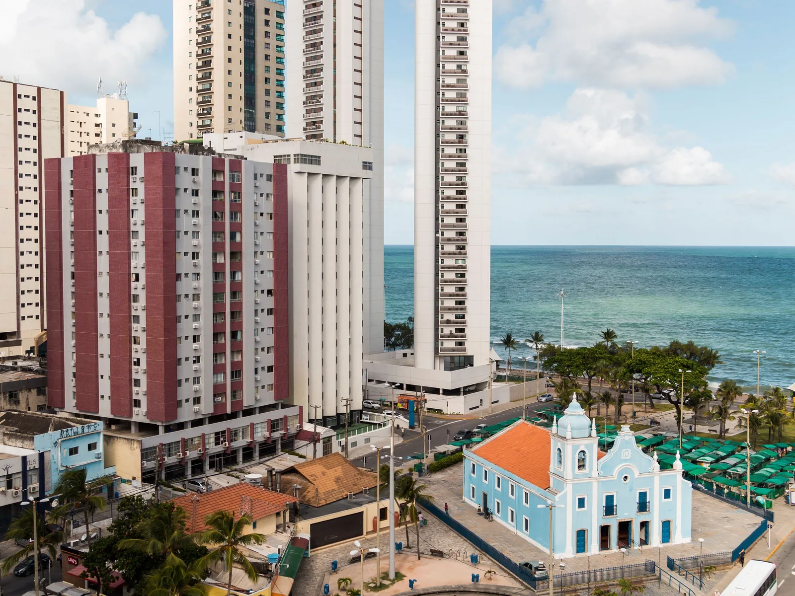 Boa Viagem from above: apartment towers crowded up to the seafront avenue, the pale blue and white church of Nossa Senhora da Boa Viagem beside the sand, and a band of shallow green water inshore of the line where the surf breaks on the reef