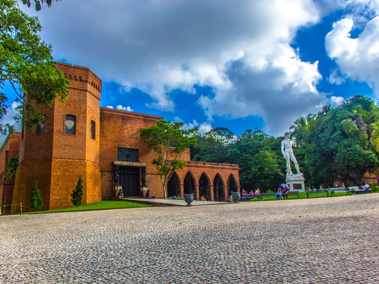 The red-brick neo-Gothic castle of the Instituto Ricardo Brennand behind a wide cobbled forecourt, its arcaded wing running away to one side under a bright sky