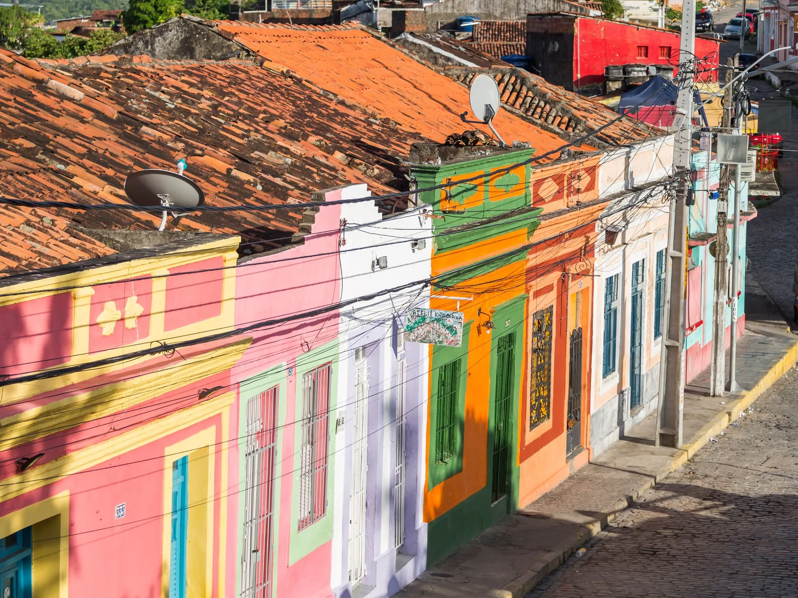 A steep cobbled lane in Olinda climbing between colonial houses painted pink, green, yellow and orange, with terracotta roofs and power lines overhead