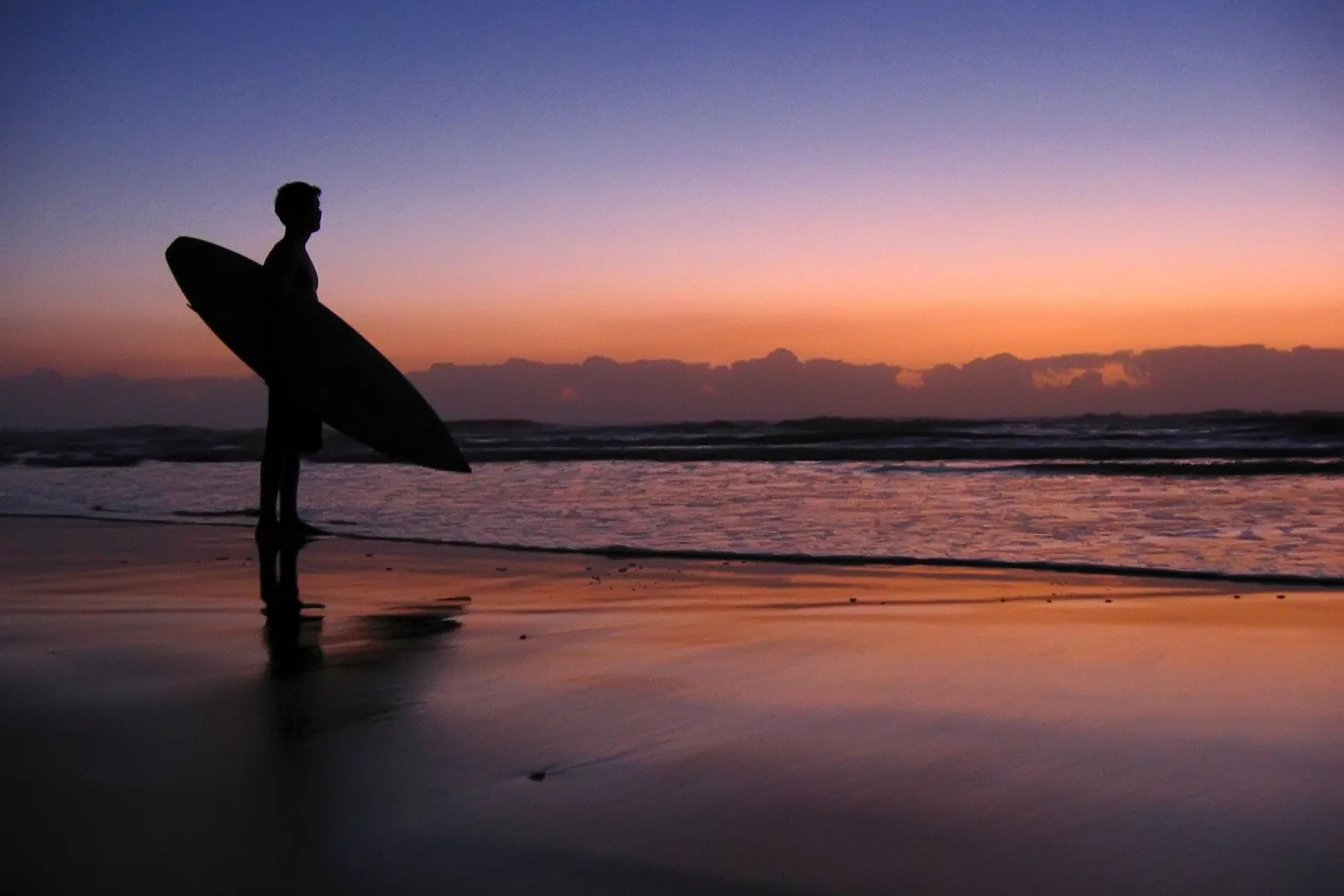 A surfer carrying a board out of the shallows at first light, the beach empty behind them