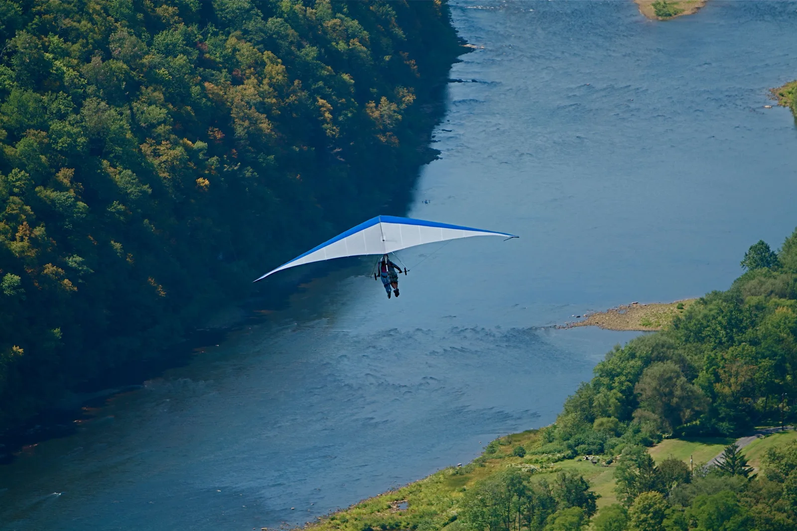 A tandem hang glider in flight high above forested slopes running down to the water