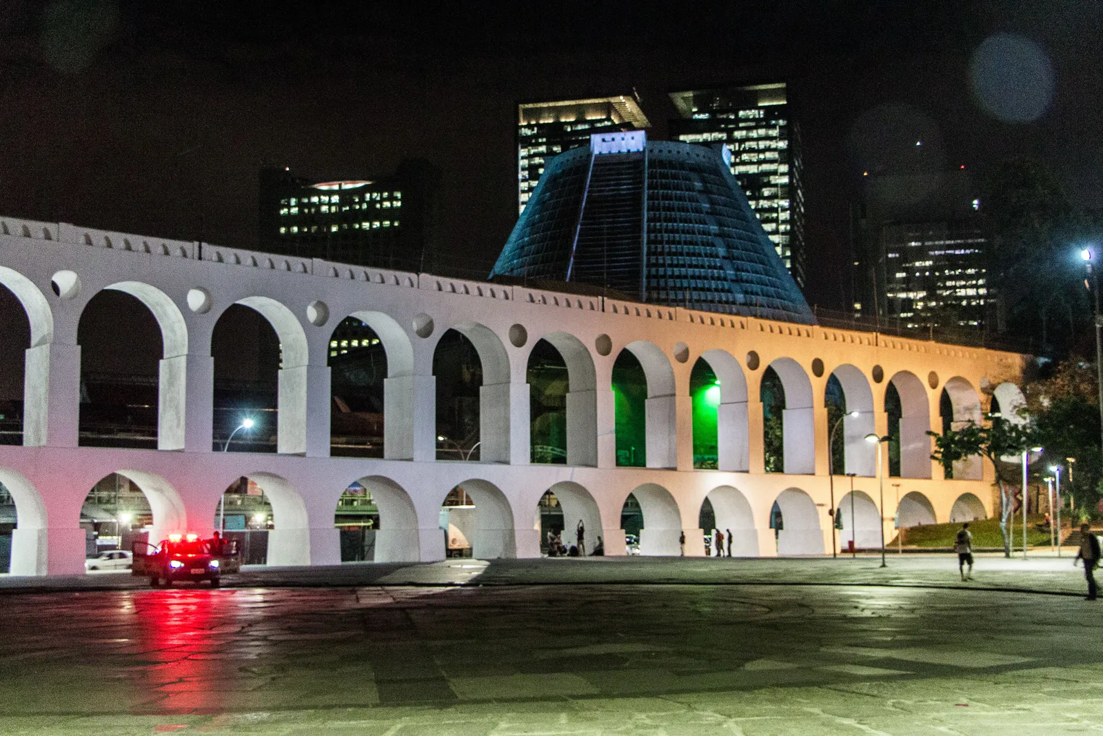 The white arches of the Lapa aqueduct floodlit at night, traffic passing beneath and the lit towers of Centro behind