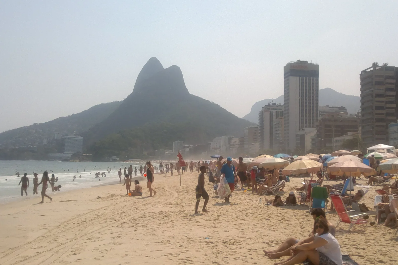 Leblon beach with people on the sand and under umbrellas, the Dois Irmãos peaks rising close at the western end