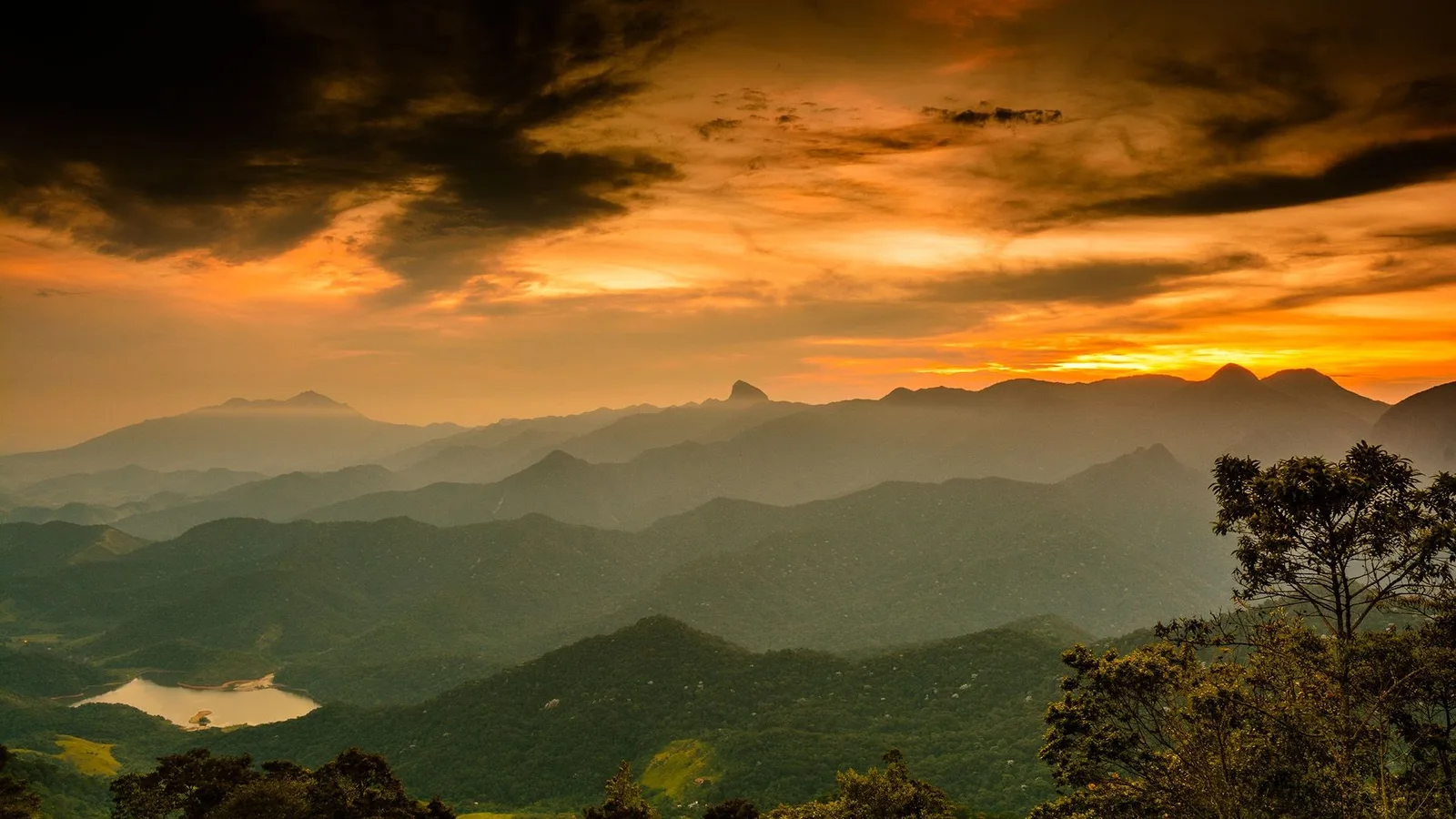 Rio at dusk from high in the hills, forested ridges falling away toward a lit coastline under a burning sky