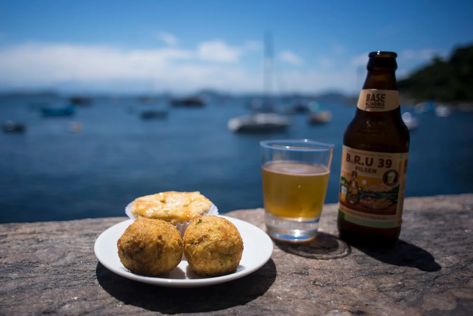 A plate of fried bolinhos and a cold beer on a stone wall above the bay, small boats moored in the water beyond