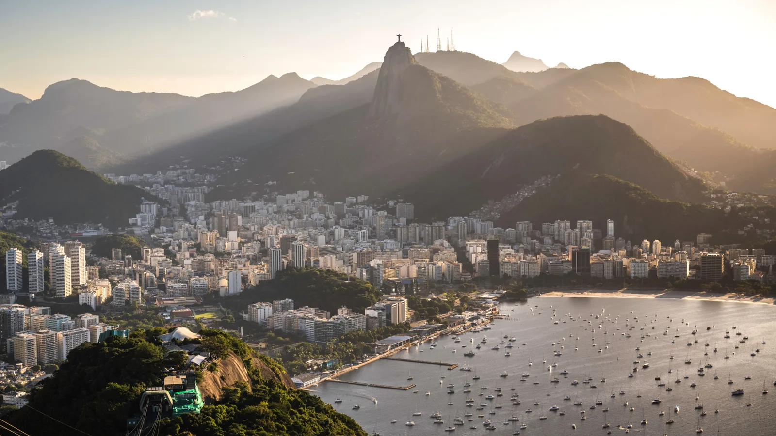 The view from Sugarloaf at sunset over Botafogo bay: yachts moored in the marina, the city stretching back between forested peaks, and Christ the Redeemer small on the Corcovado ridge above it