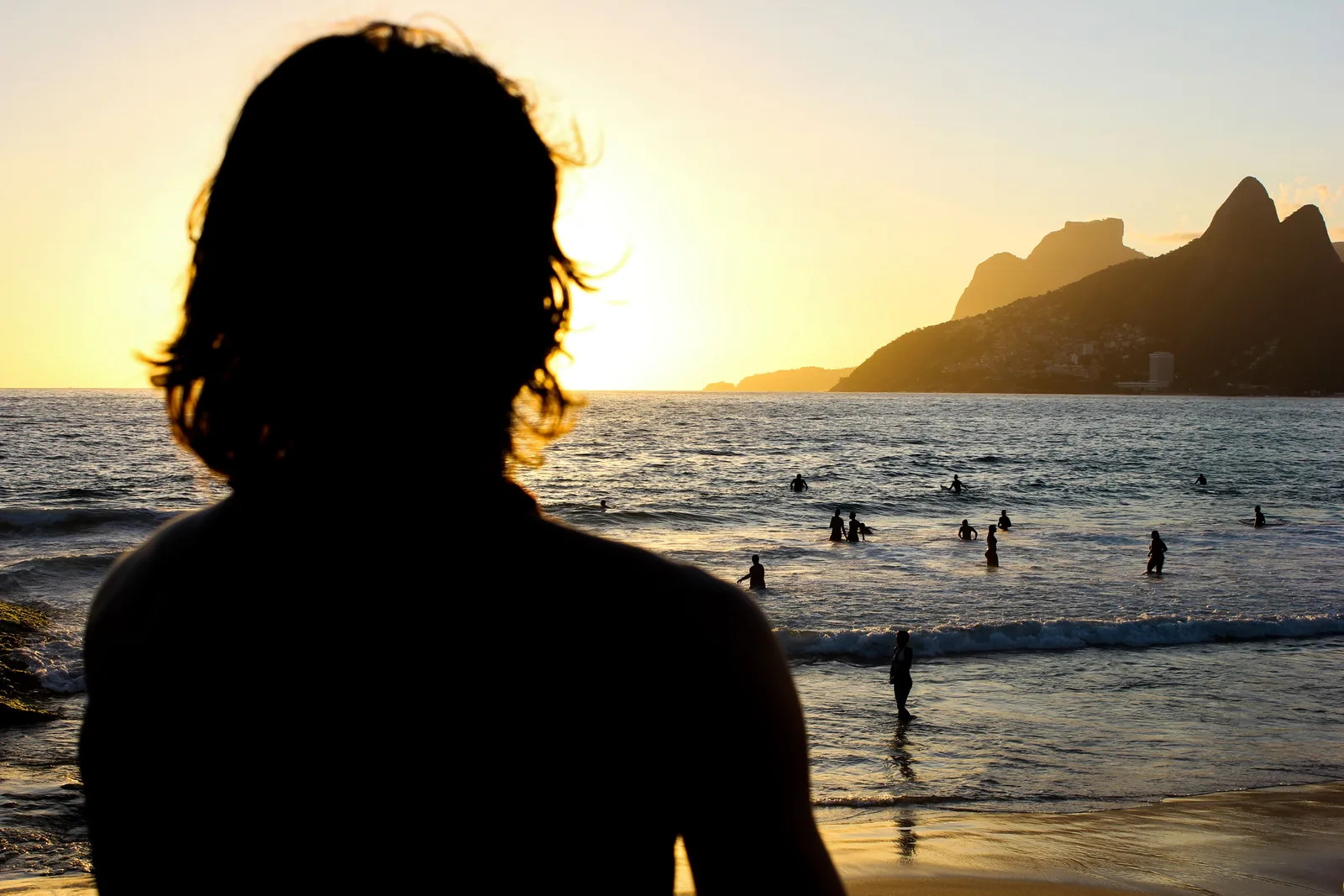 A figure silhouetted against the sunset at Arpoador, swimmers still in the water and the Dois Irmãos peaks dark at the end of the beach