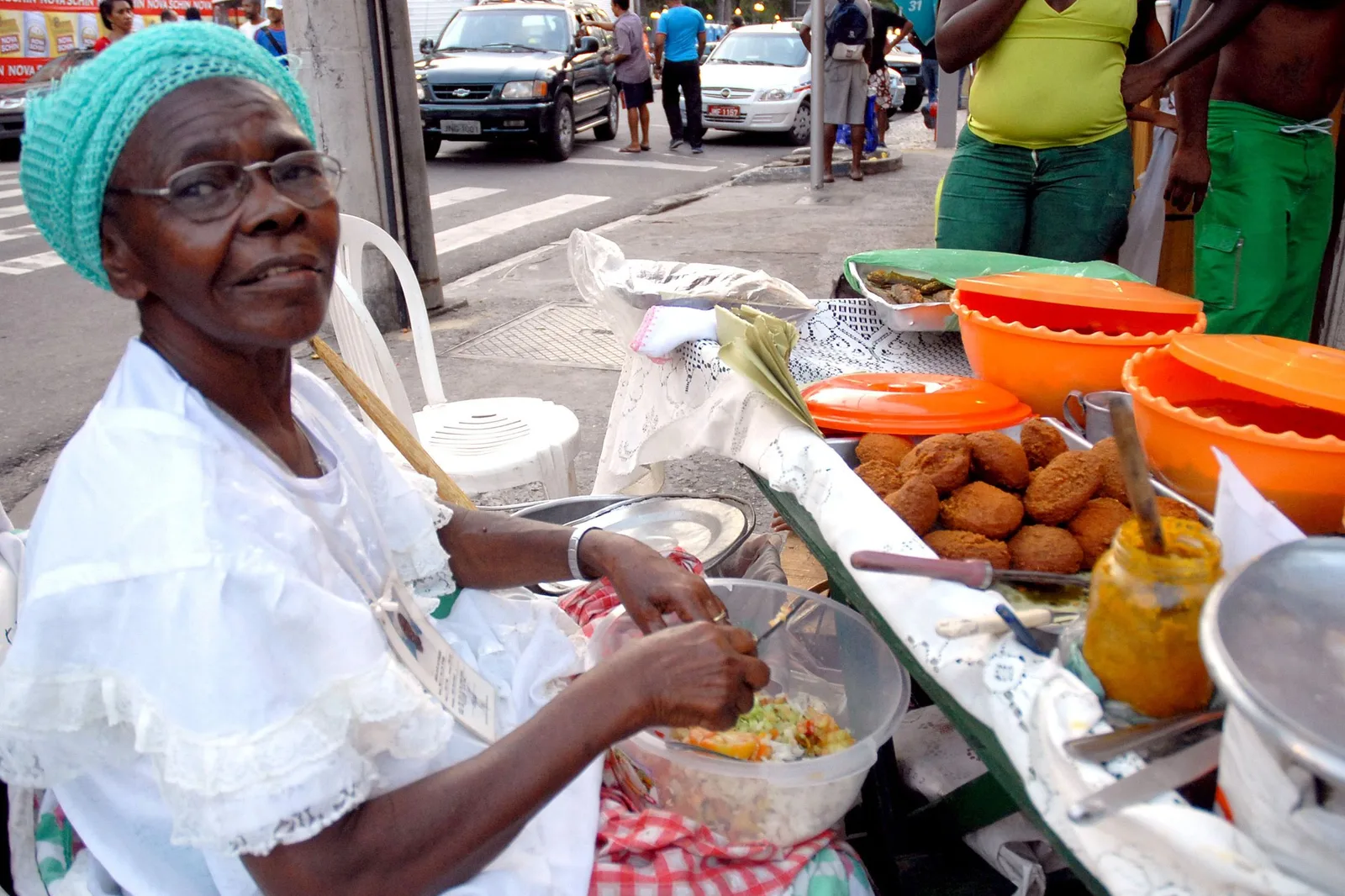 A baiana in white lace and a knitted headwrap working at her street acarajé stand, a tray of fried fritters and covered orange bowls of fillings laid out along the table beside her