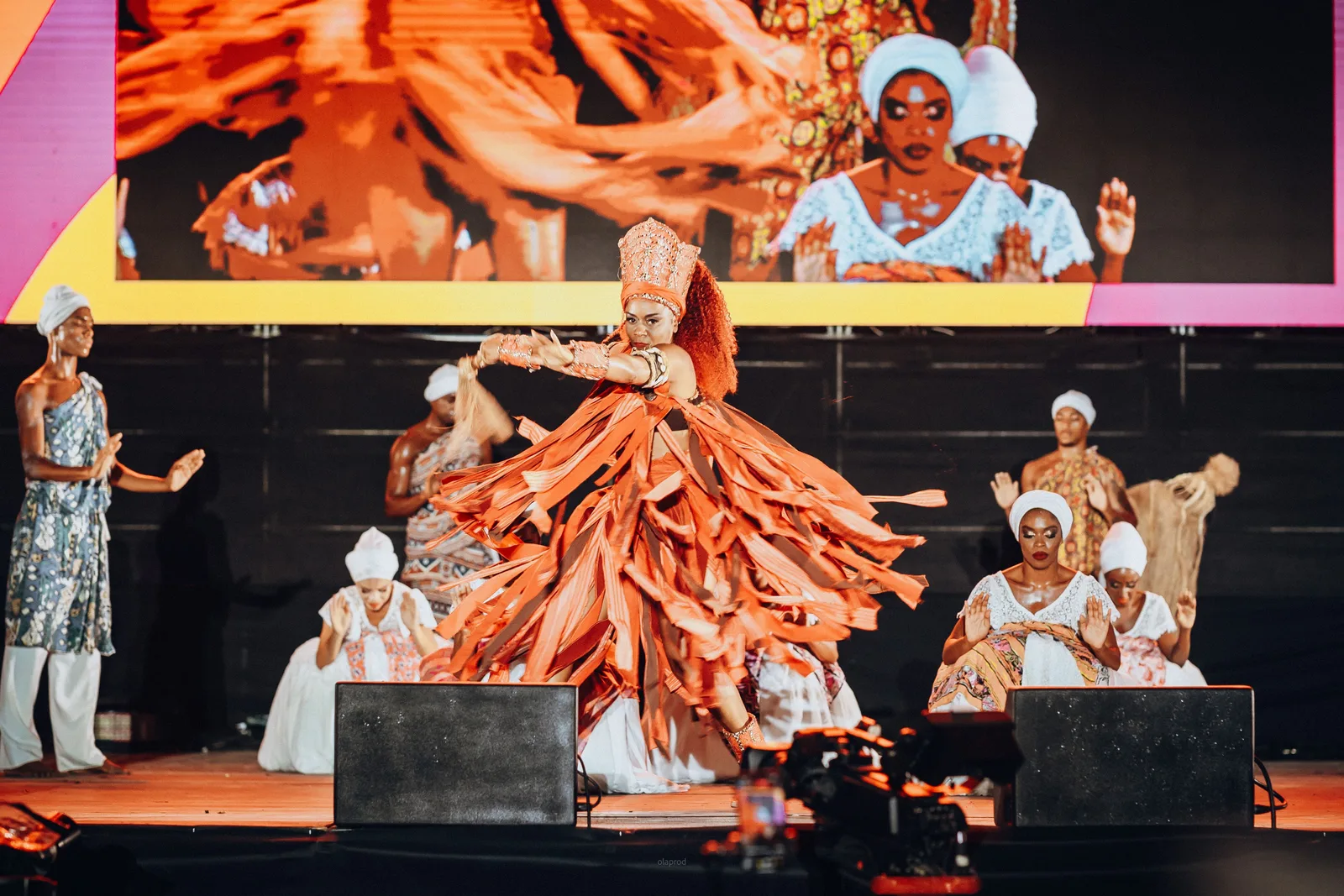 A dancer of the Balé Folclórico da Bahia mid-spin in a full orange raffia orixá costume and beaded crown, the rest of the company kneeling in white on the darkened stage behind her