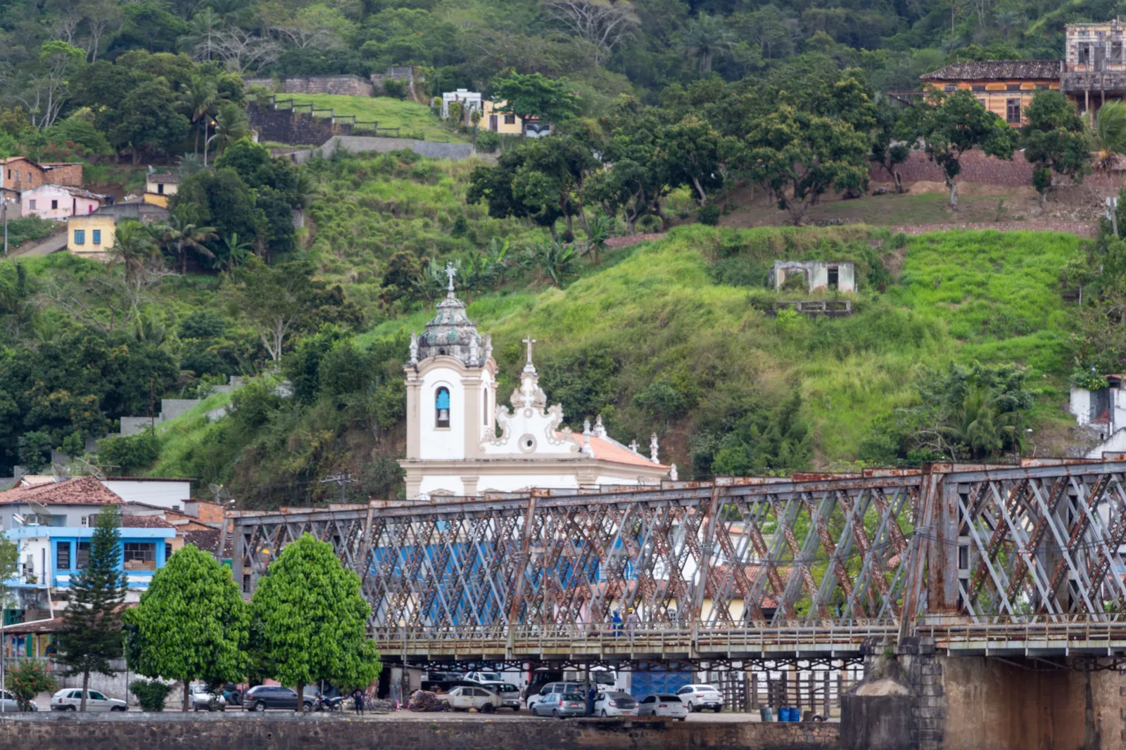 The iron Dom Pedro II bridge between Cachoeira and São Félix, its rusting lattice spans crossing the frame in front of a white baroque church tower and the pastel houses climbing the green hillside behind